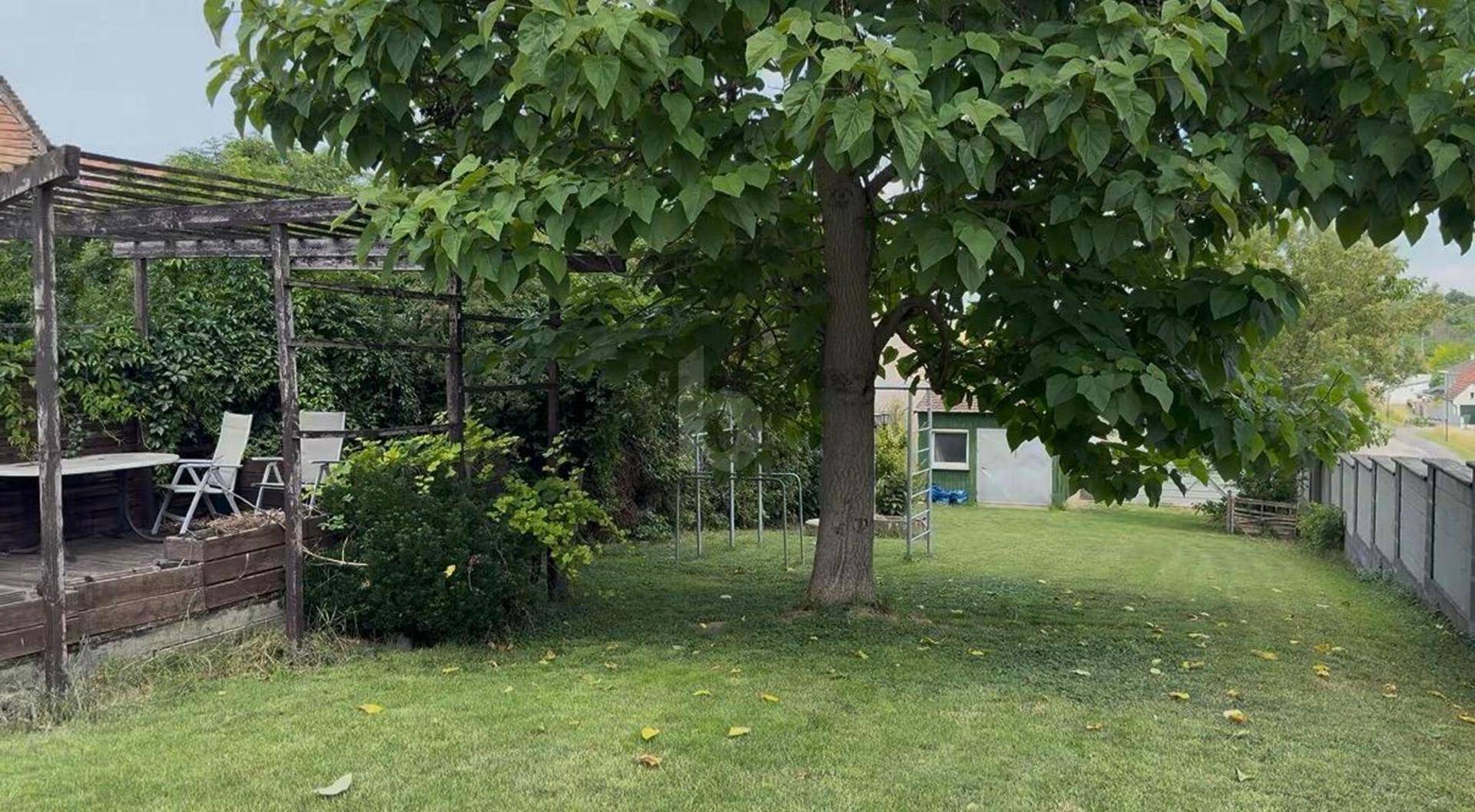 Großer Garten mit altem Baum, Rasenfläche und einer Holzpergola mit Sitzbereich.