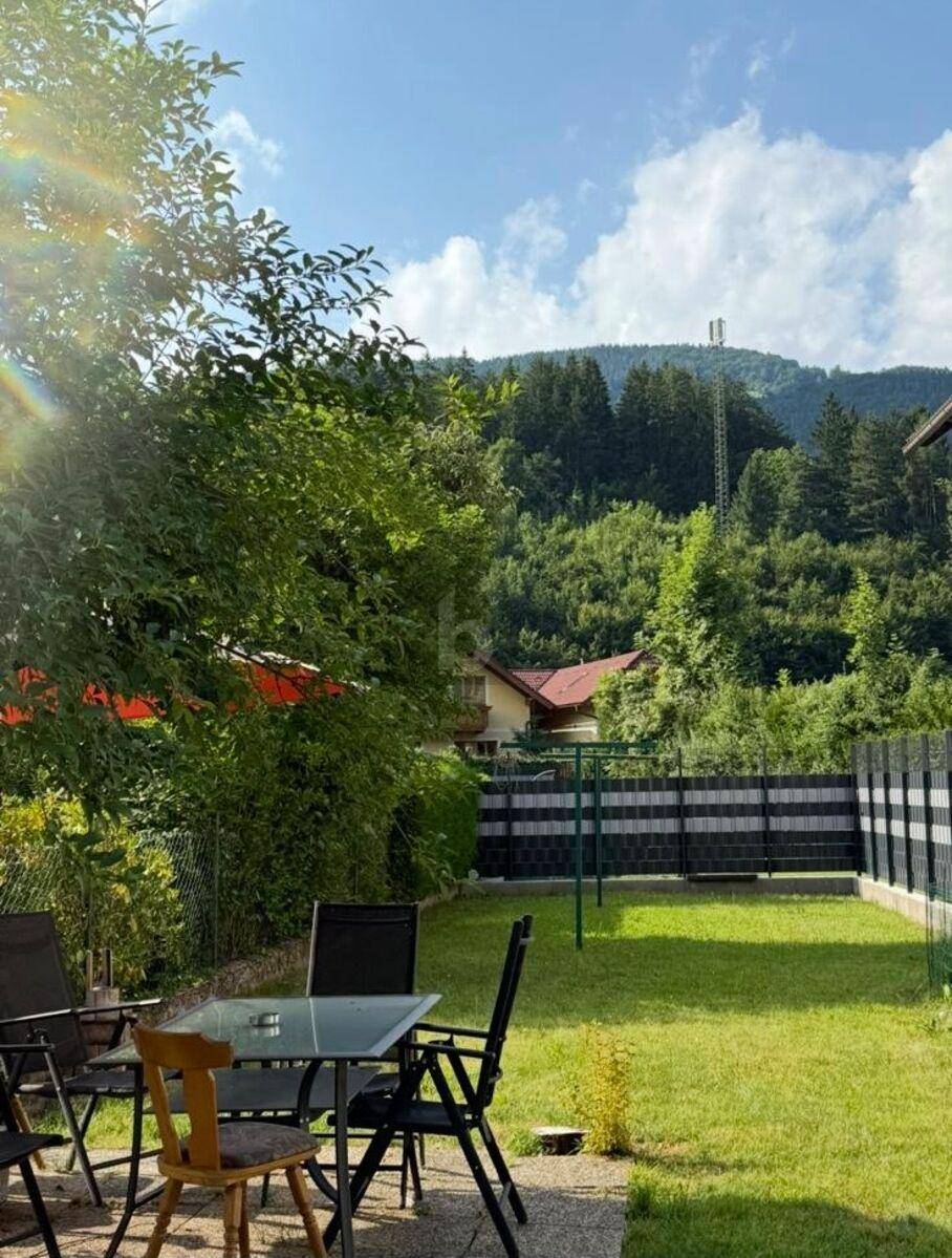 Sonniger Garten mit Terrasse, Sitzgelegenheiten und Blick auf bewaldete Hügel.