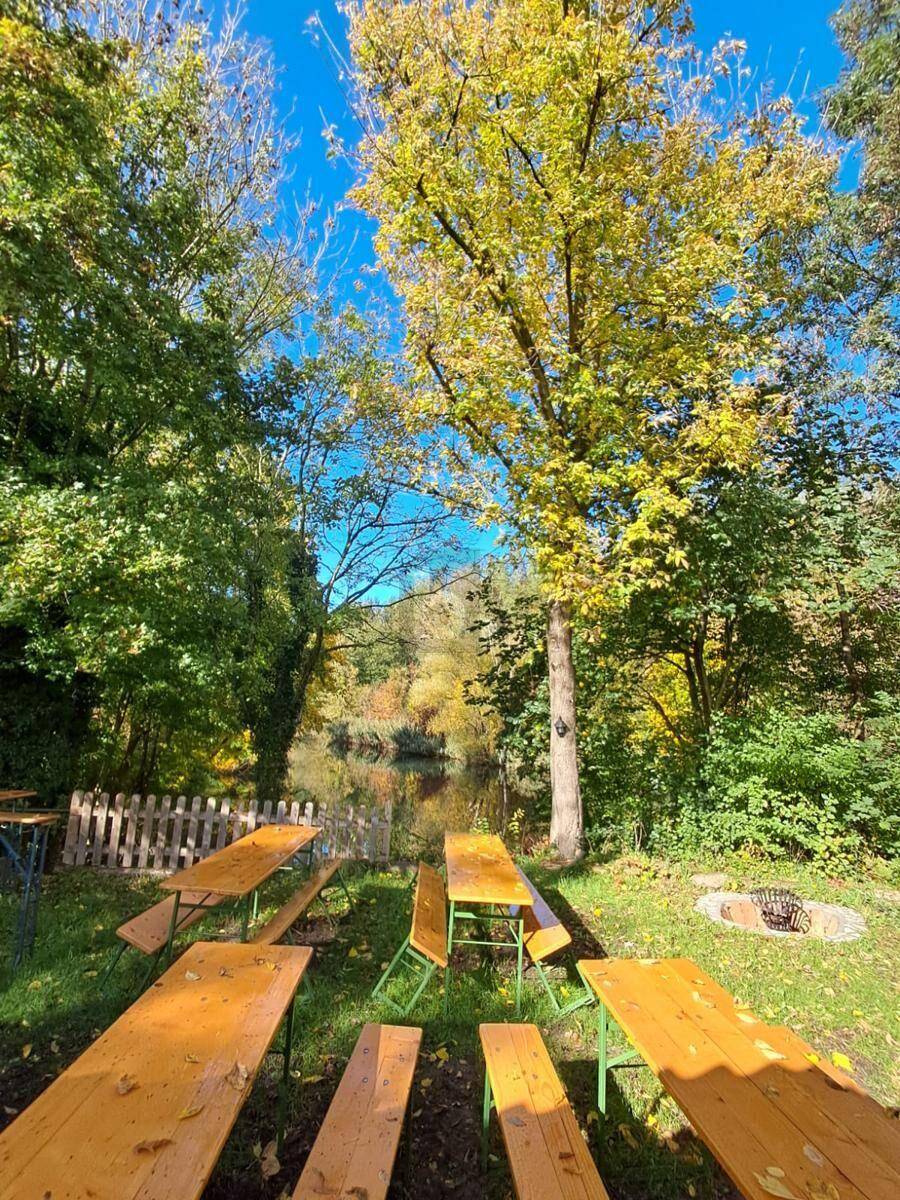 Gemütlicher Außenbereich mit Holztischen und Bänken direkt am Wasser, umgeben von herbstlichen Bäumen.