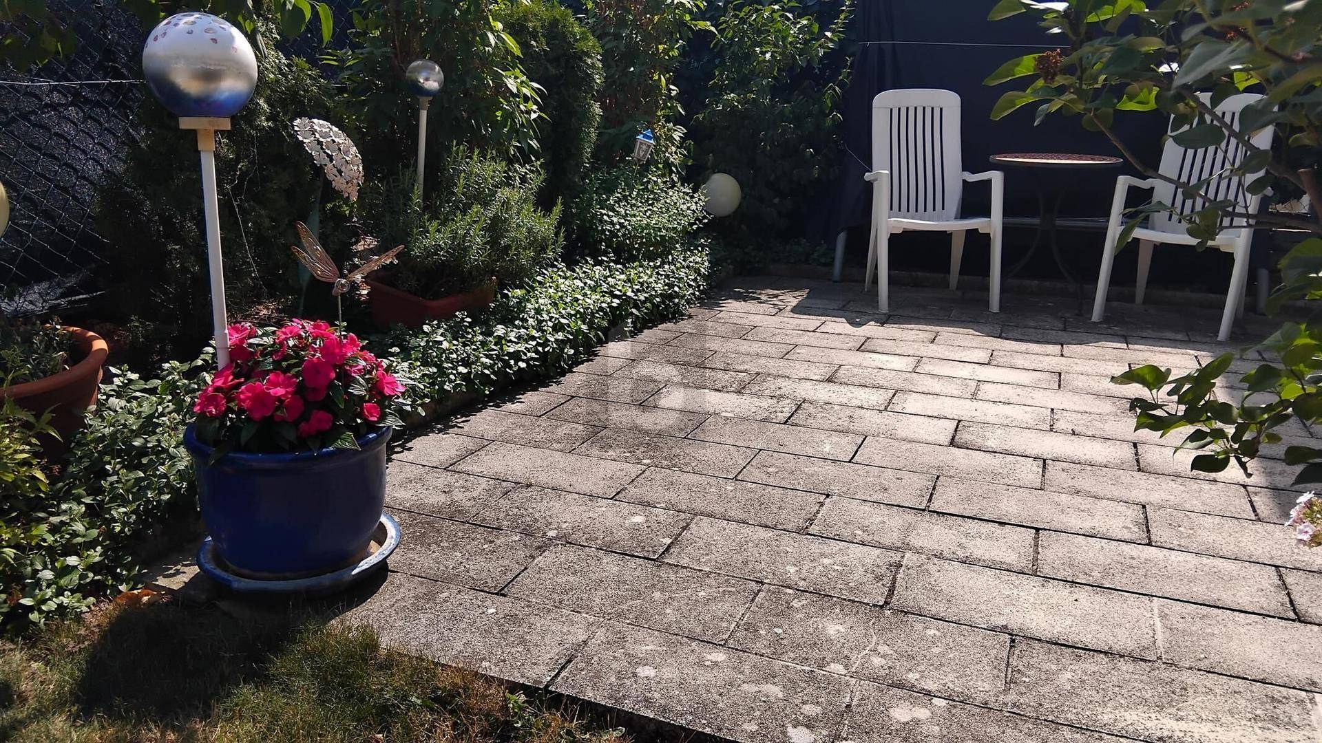 Gepflasterte Terrasse im Garten mit Blumenkübeln und Gartenmöbeln, umgeben von grünen Pflanzen.
