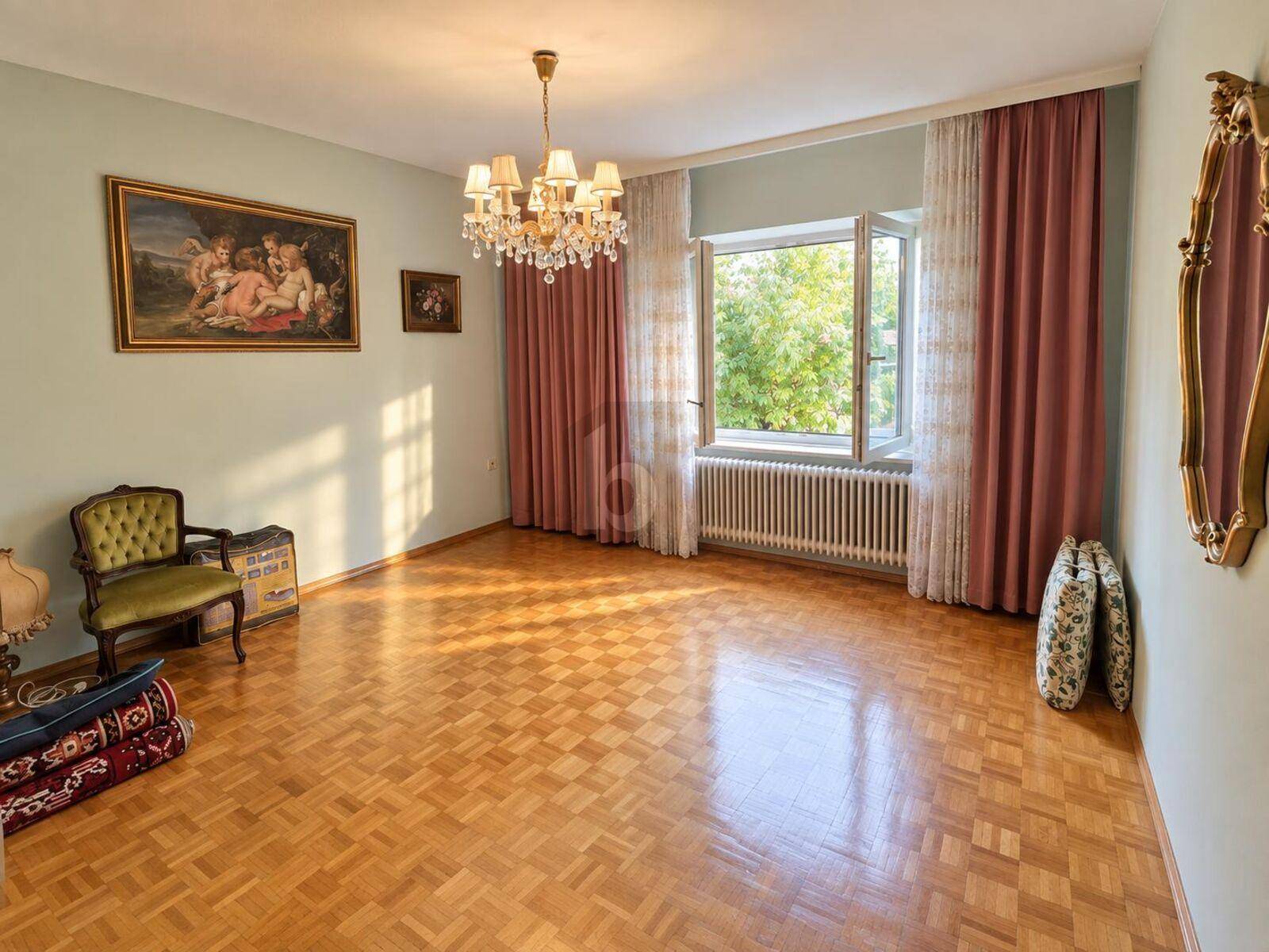 Helles Wohnzimmer mit Fischgrätparkett, einem Kronleuchter und großen Fenstern mit Blick ins Grüne.
