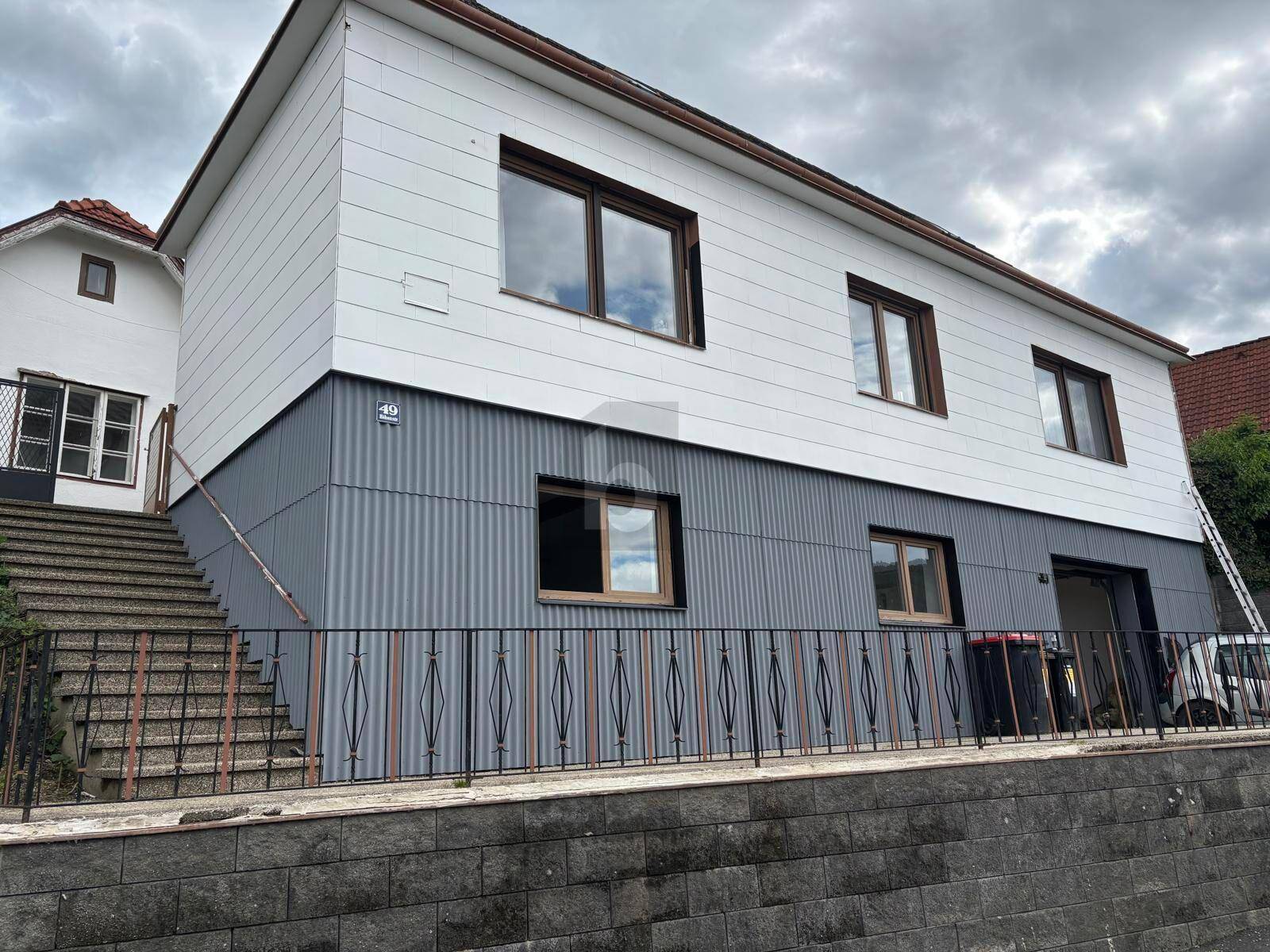 Fassade eines Hauses mit grauer Wellblechverkleidung im Erdgeschoss und weißer Holzverkleidung im Obergeschoss.