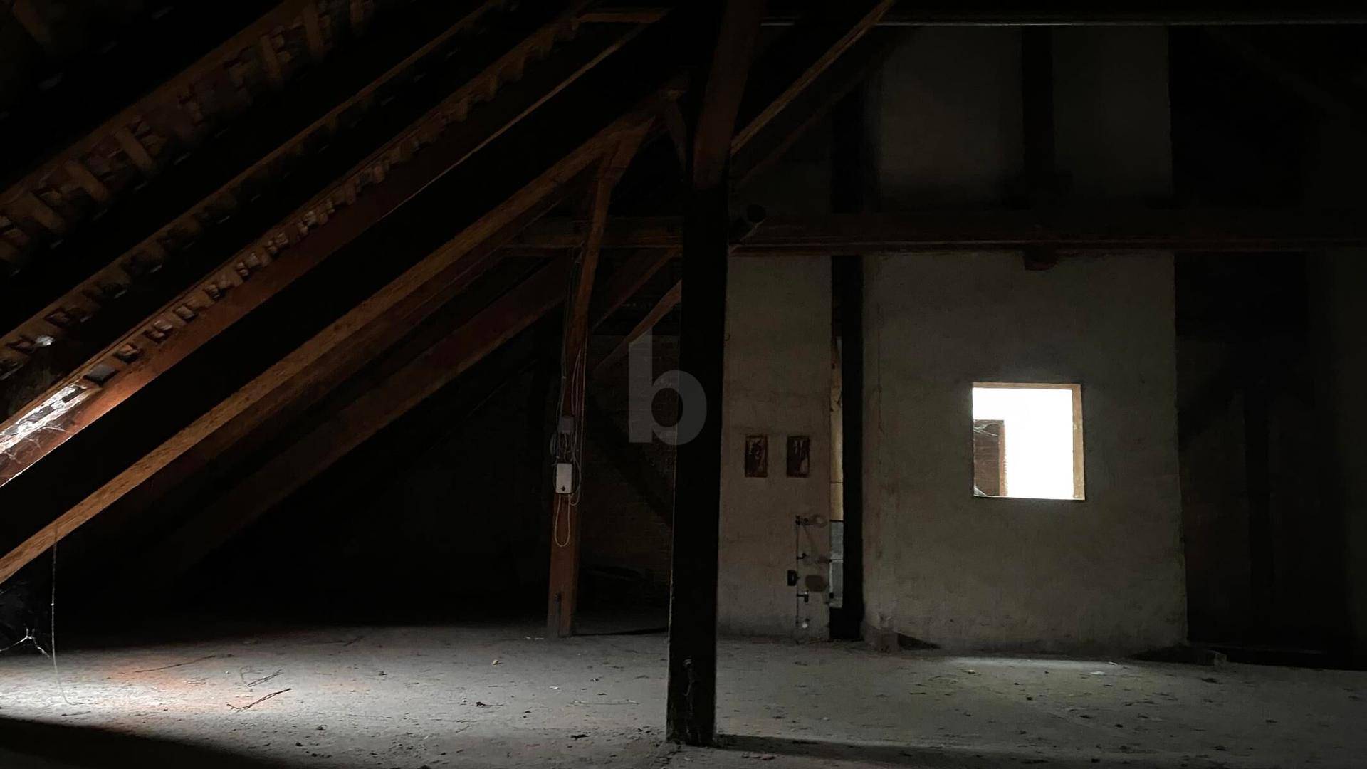 Dunkler Dachboden mit sichtbaren Holzbalken und einem kleinen Fenster, das Licht hereinlässt.