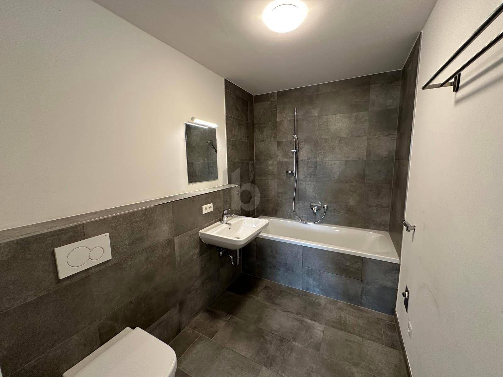 Modernes Badezimmer mit grauen Fliesen, Badewanne mit Duschfunktion und wandhängendem WC.