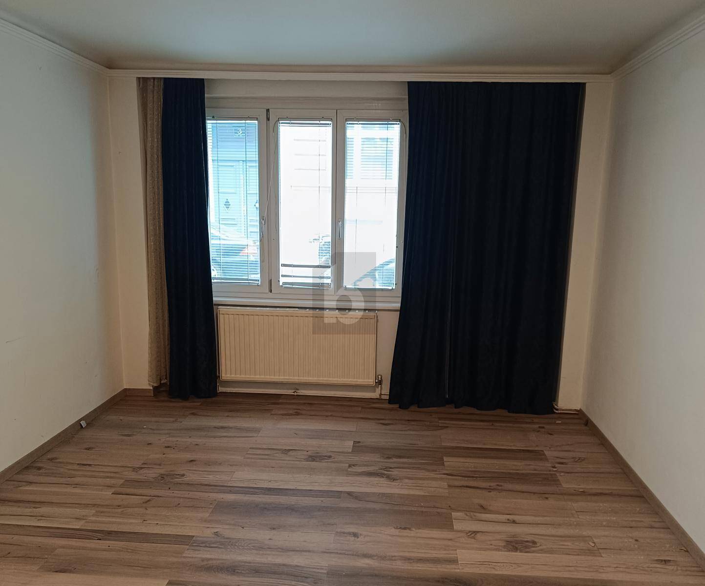 Leerer Raum mit Holzoptikboden, weißen Wänden und Fenster mit dunklen Vorhängen.