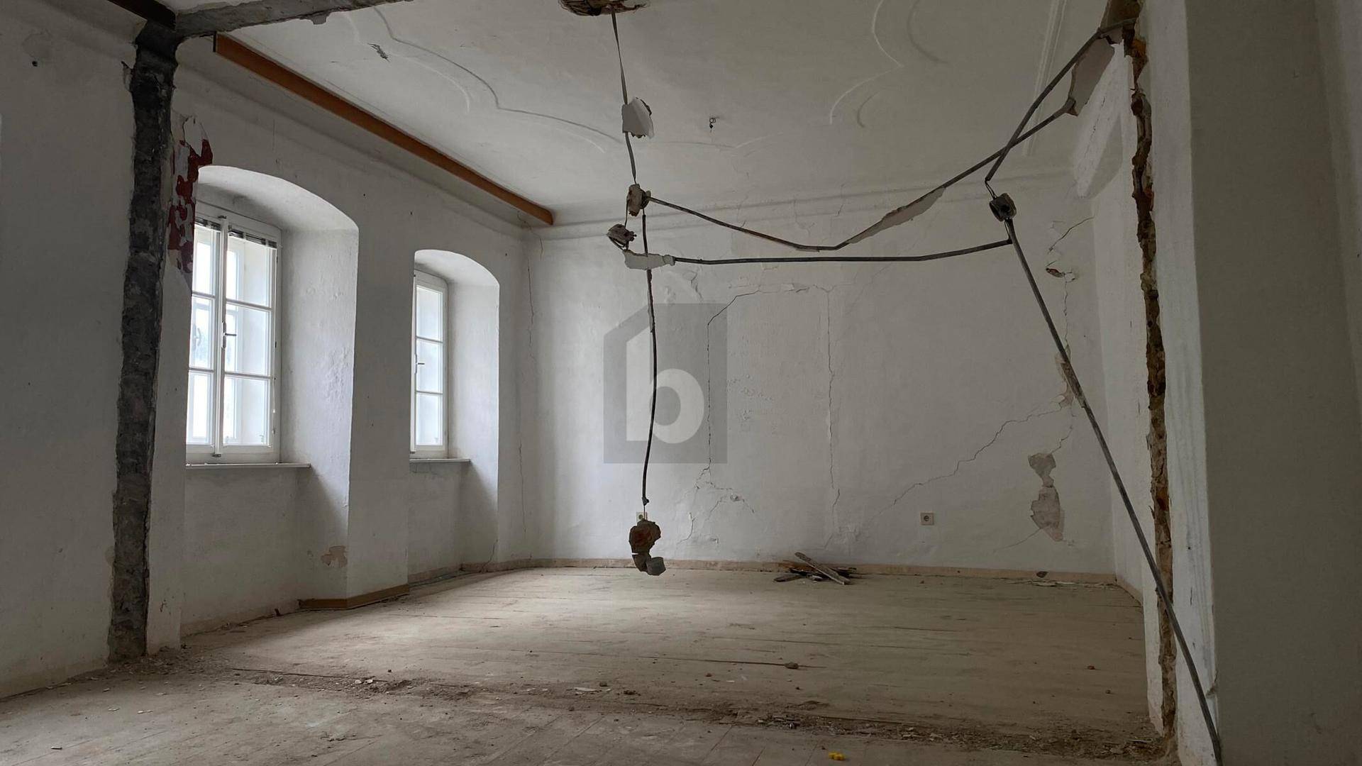 Leerer Raum unter Renovierung mit freigelegten Wänden, alten Fenstern und Stuckdecke.