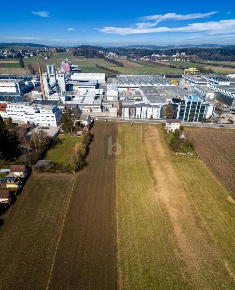 Weitläufige Luftaufnahme mit Feldern, einem Industriegebiet und hügeliger Landschaft im Hintergrund.