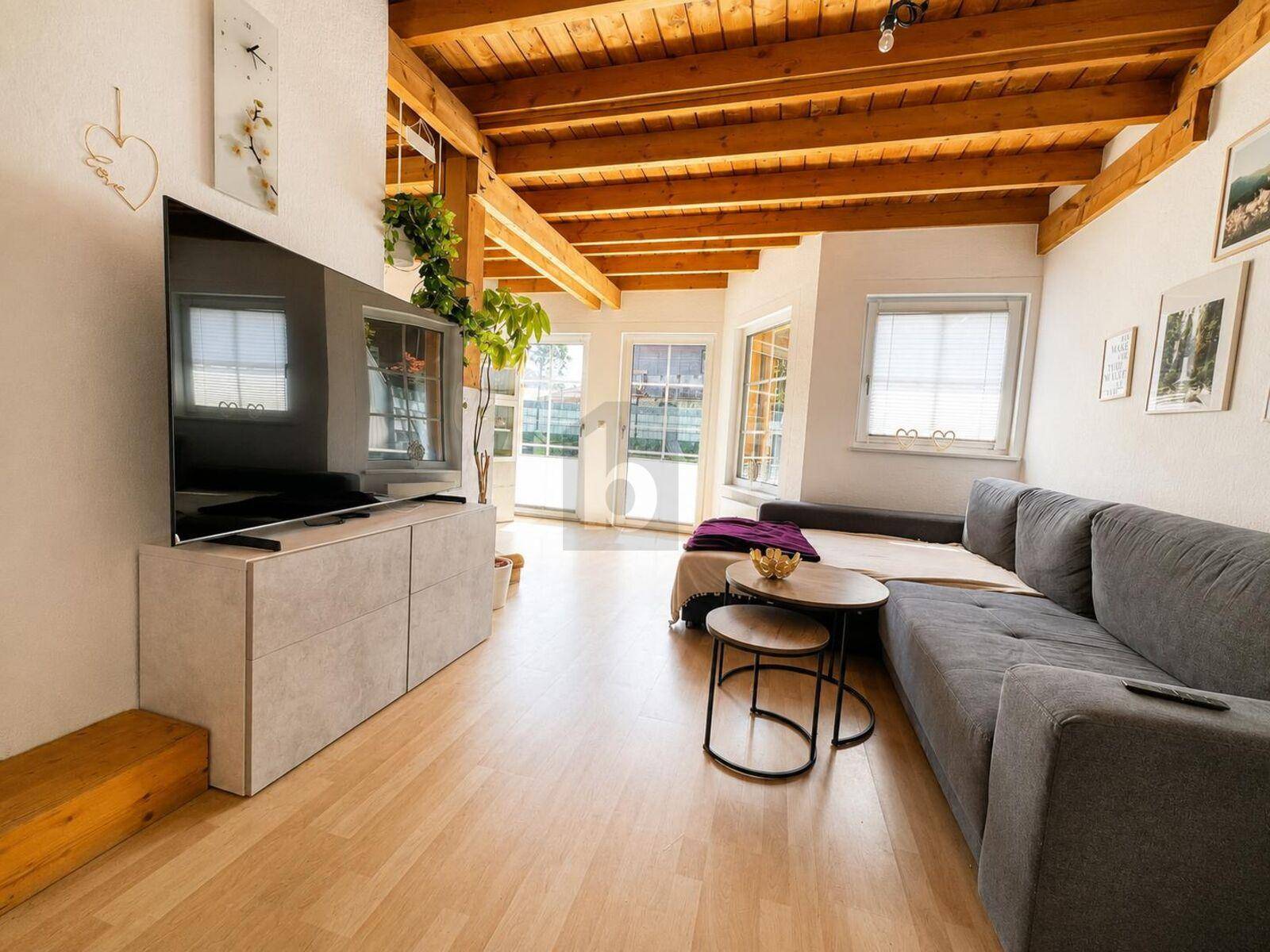 Gemütliches Wohnzimmer mit grauem Sofa, Holzboden und charakteristischer Holzdeckenkonstruktion.