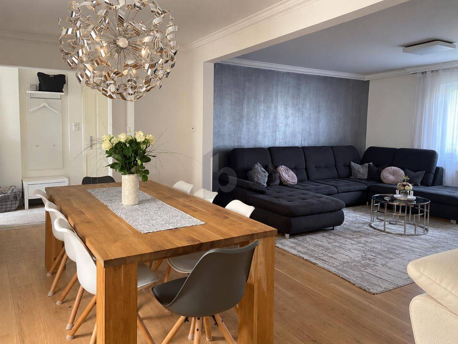 Heller offener Wohn-Essbereich mit Holztisch, bequemer Couch und modernem Kronleuchter als Blickfang.