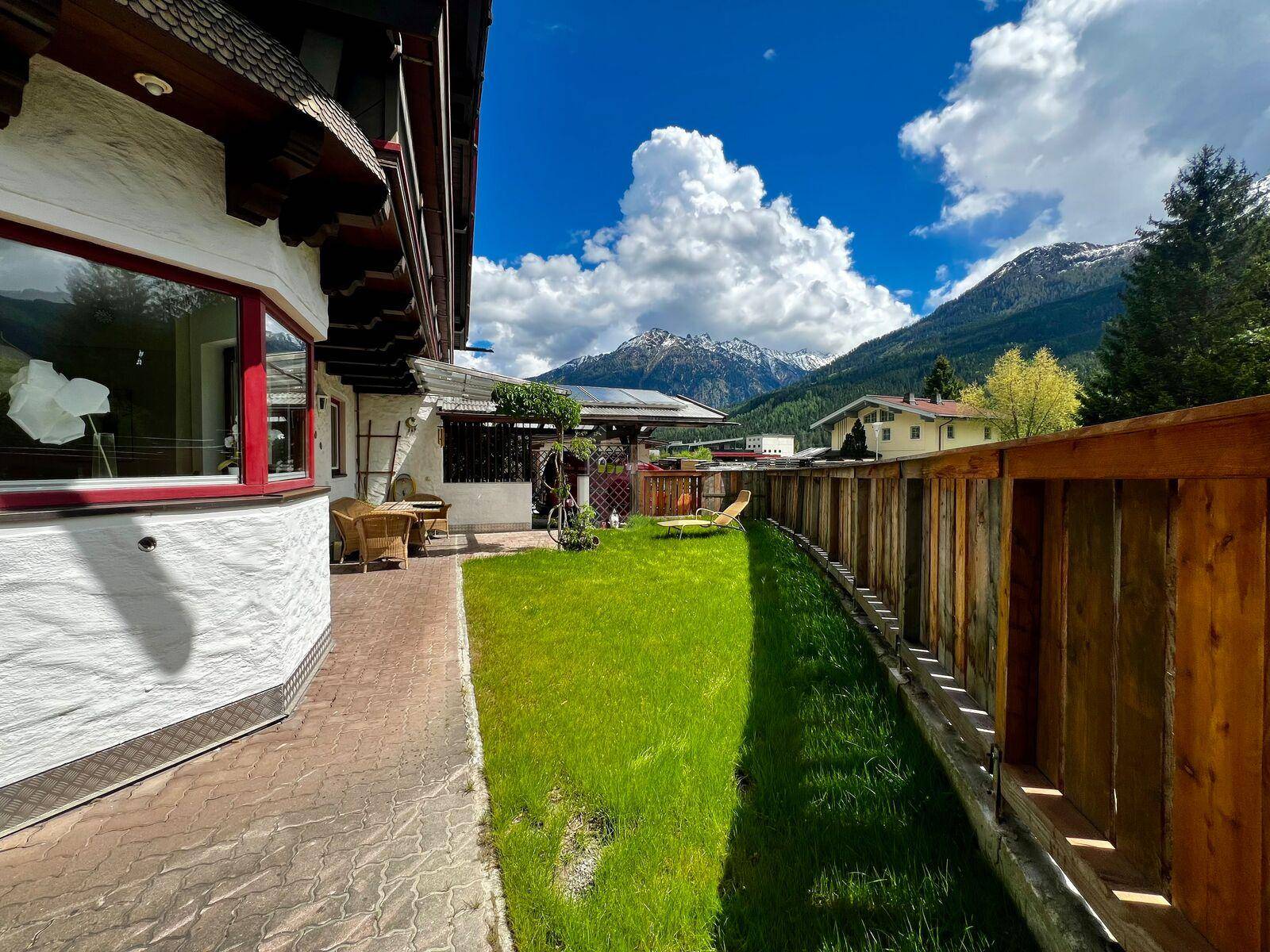 Seitlicher Blick auf das Haus mit langem Rasenbereich, Holzzaun und beeindruckendem Bergblick.