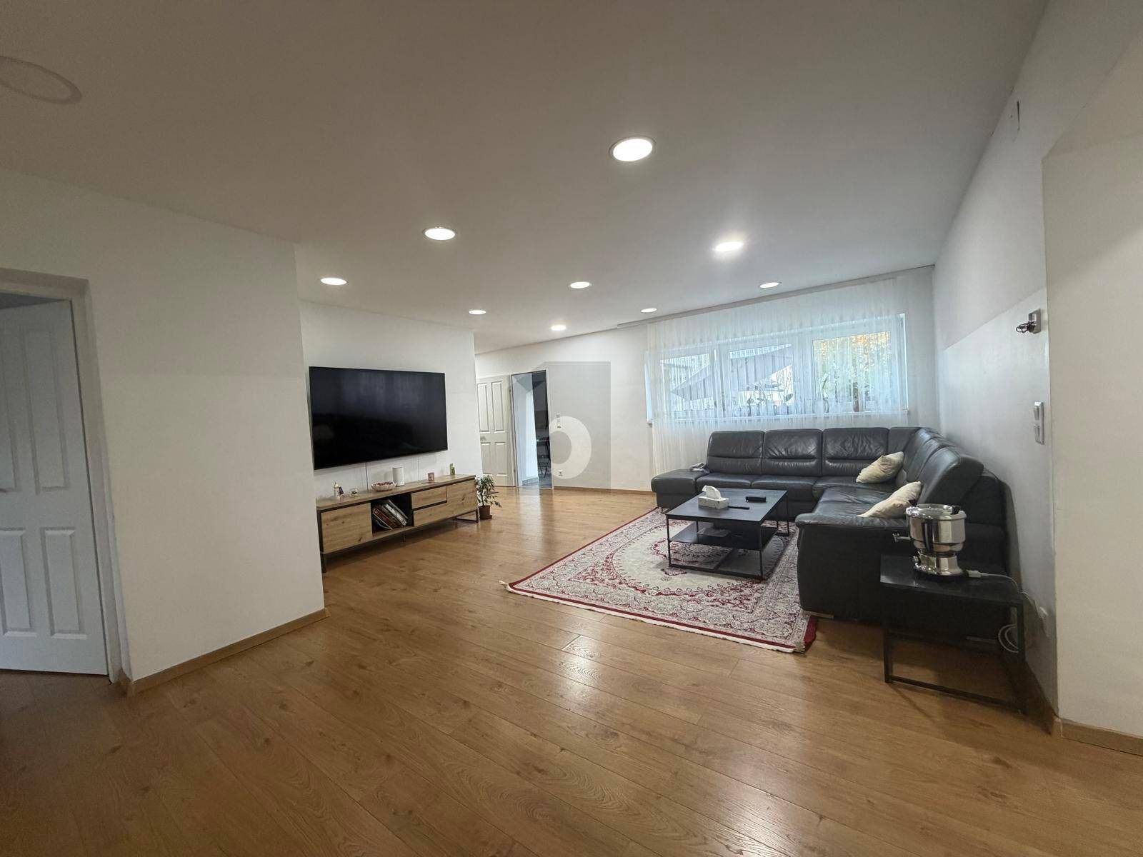 Geräumiges Wohnzimmer mit Laminatboden, großem Fernseher und viel natürlichem Licht.