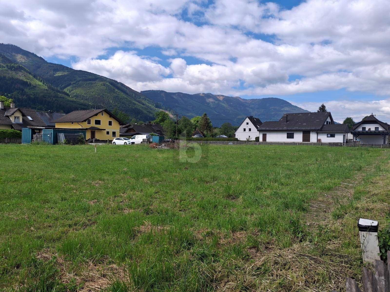Offenes Grundstück mit grüner Wiese, umgeben von ländlichen Häusern und einer weiten Berglandschaft.