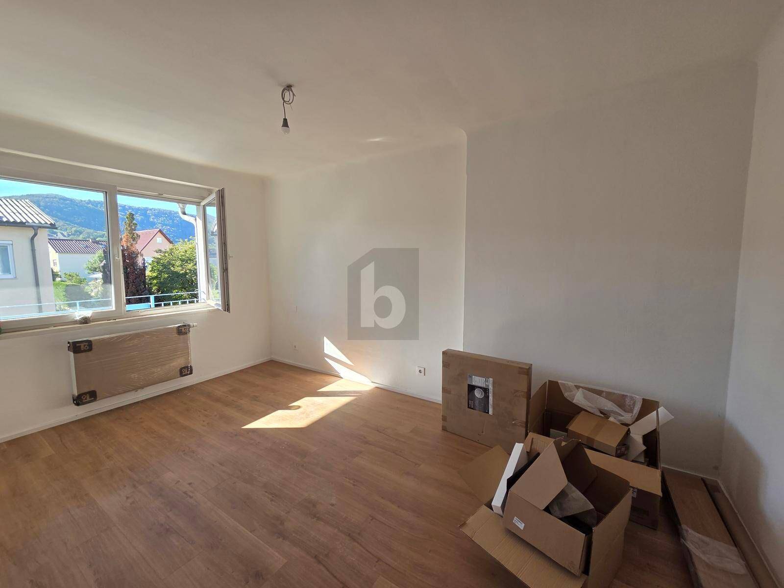 Lichtdurchflutetes Zimmer mit Laminatboden und Blick auf die umliegende Landschaft und Häuser.