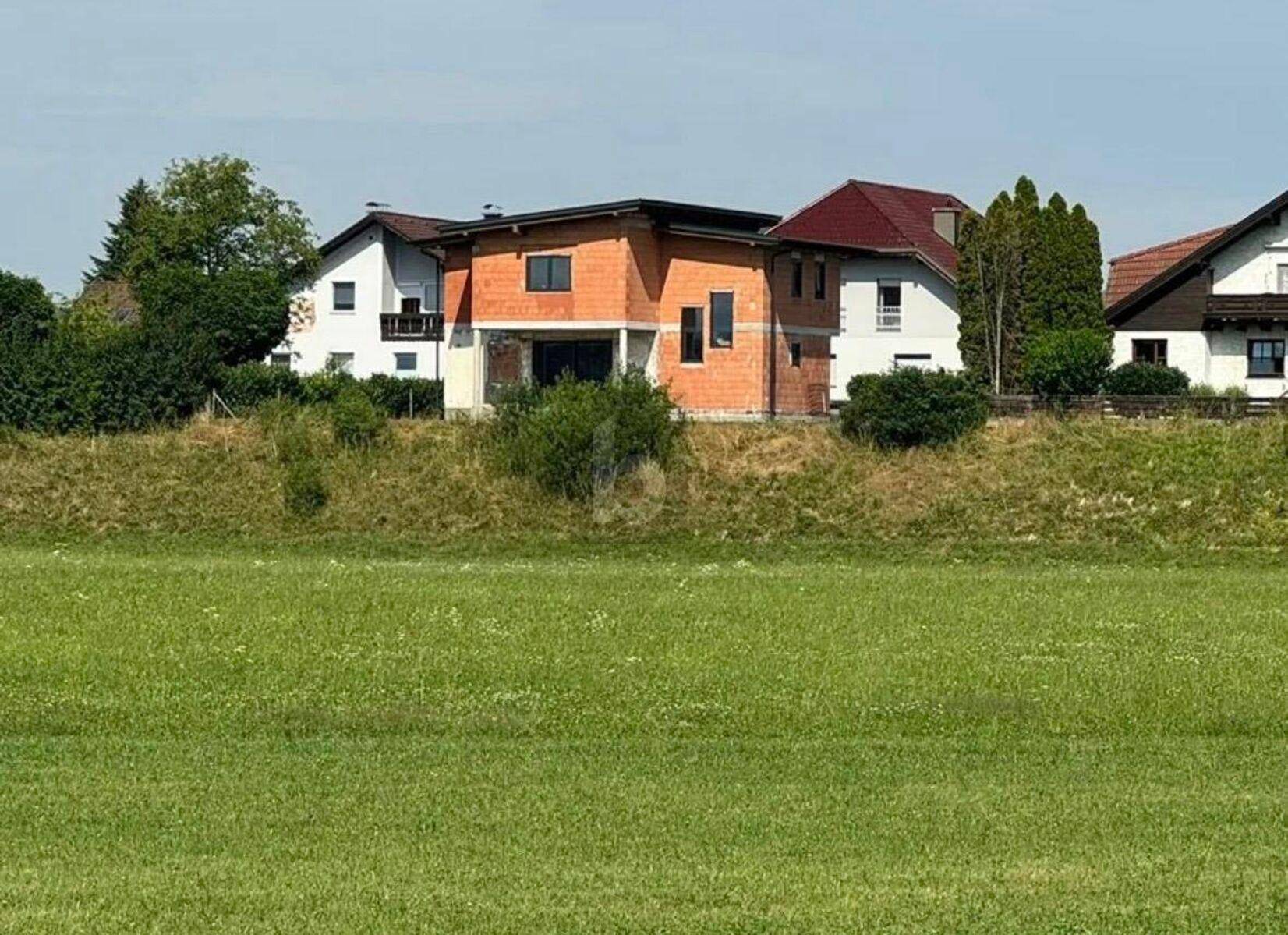 Neubau-Einfamilienhaus im Rohbau, umgeben von grünen Feldern und weiteren Wohnhäusern.