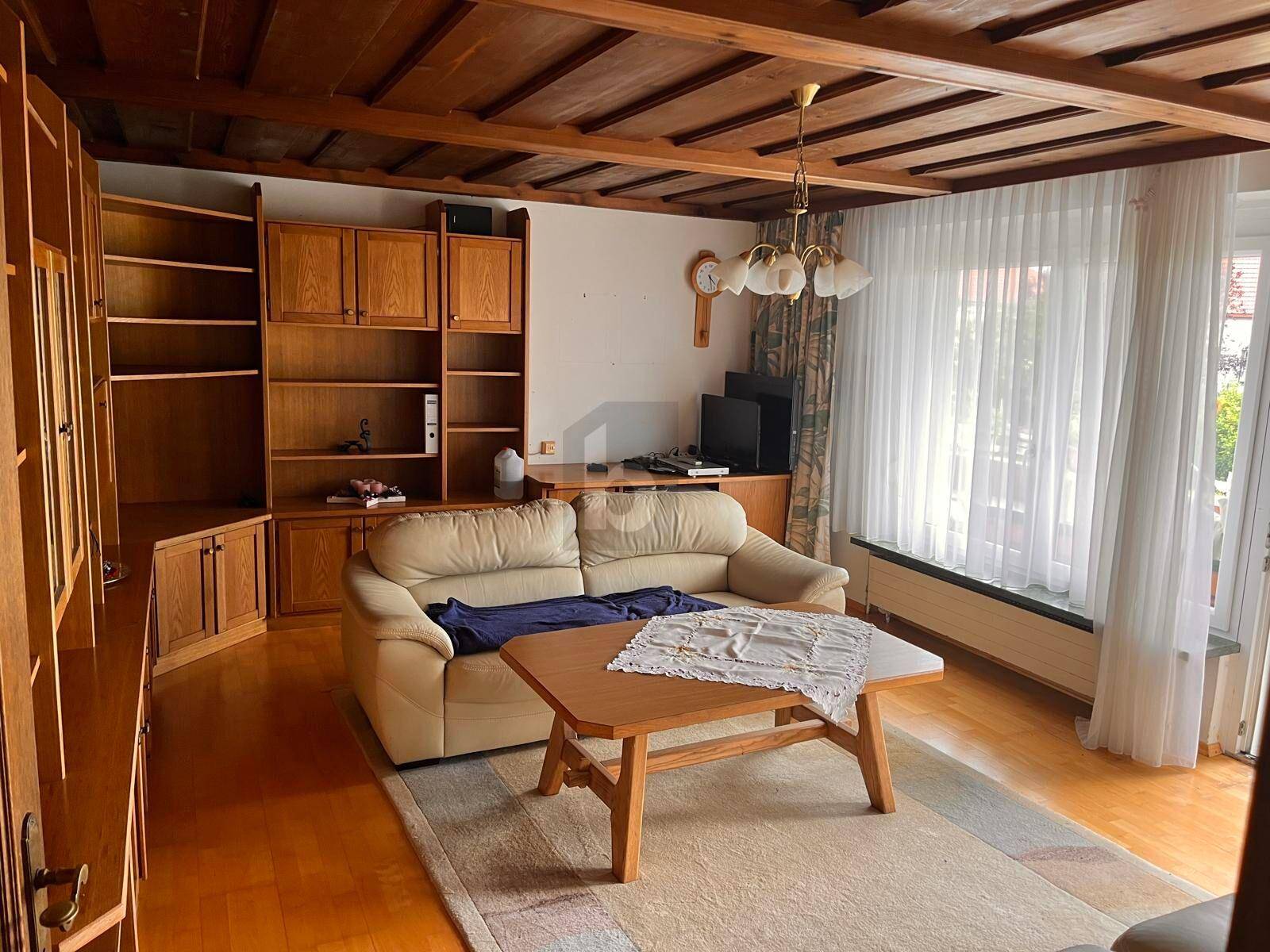 Wohnzimmer mit Holzbalkendecke, Parkettboden, beiger Couch und großem Fenster.