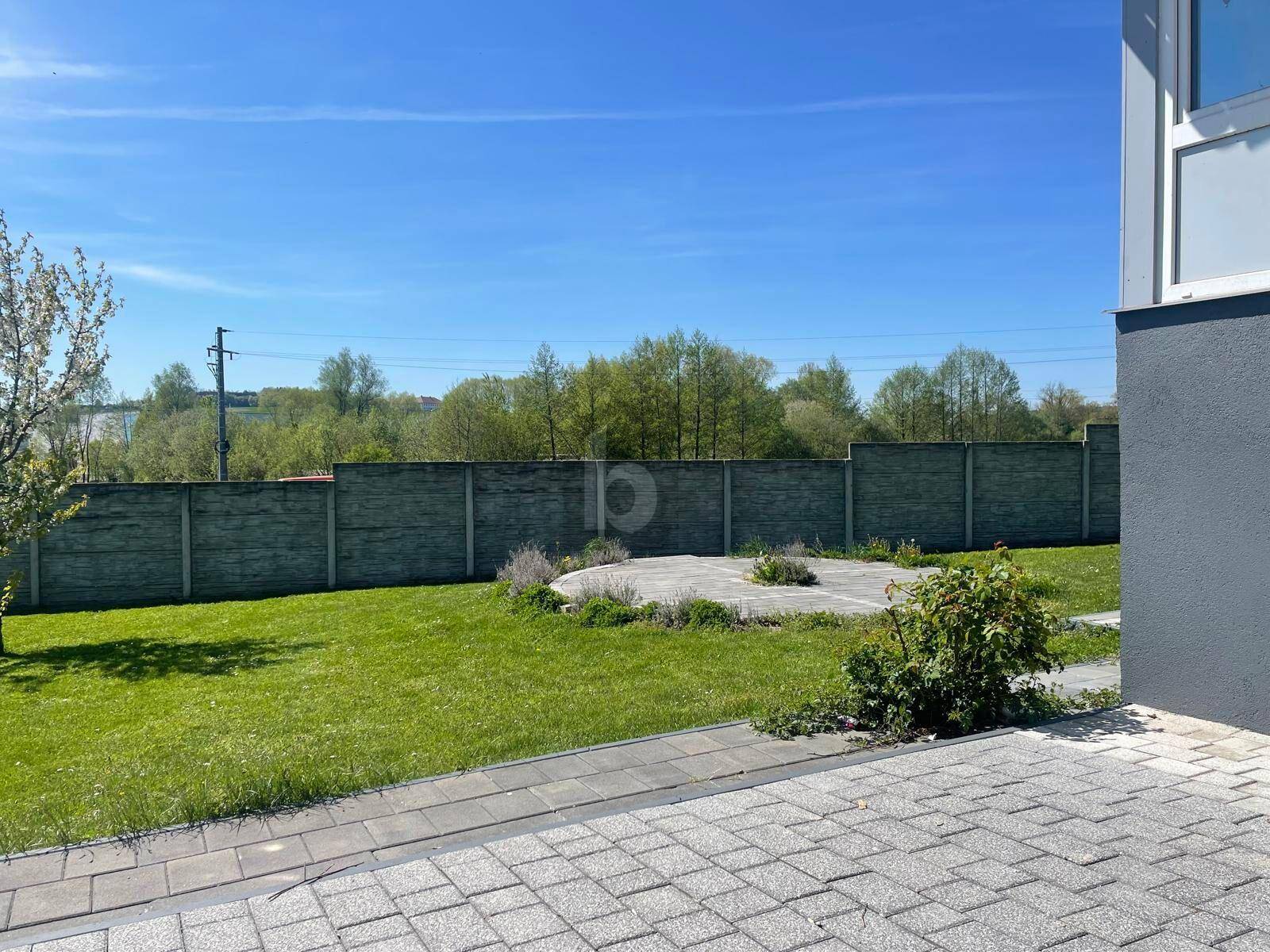 Grüner Gartenbereich mit Rasenfläche, gepflastertem Weg und einer Betonmauer im Hintergrund.