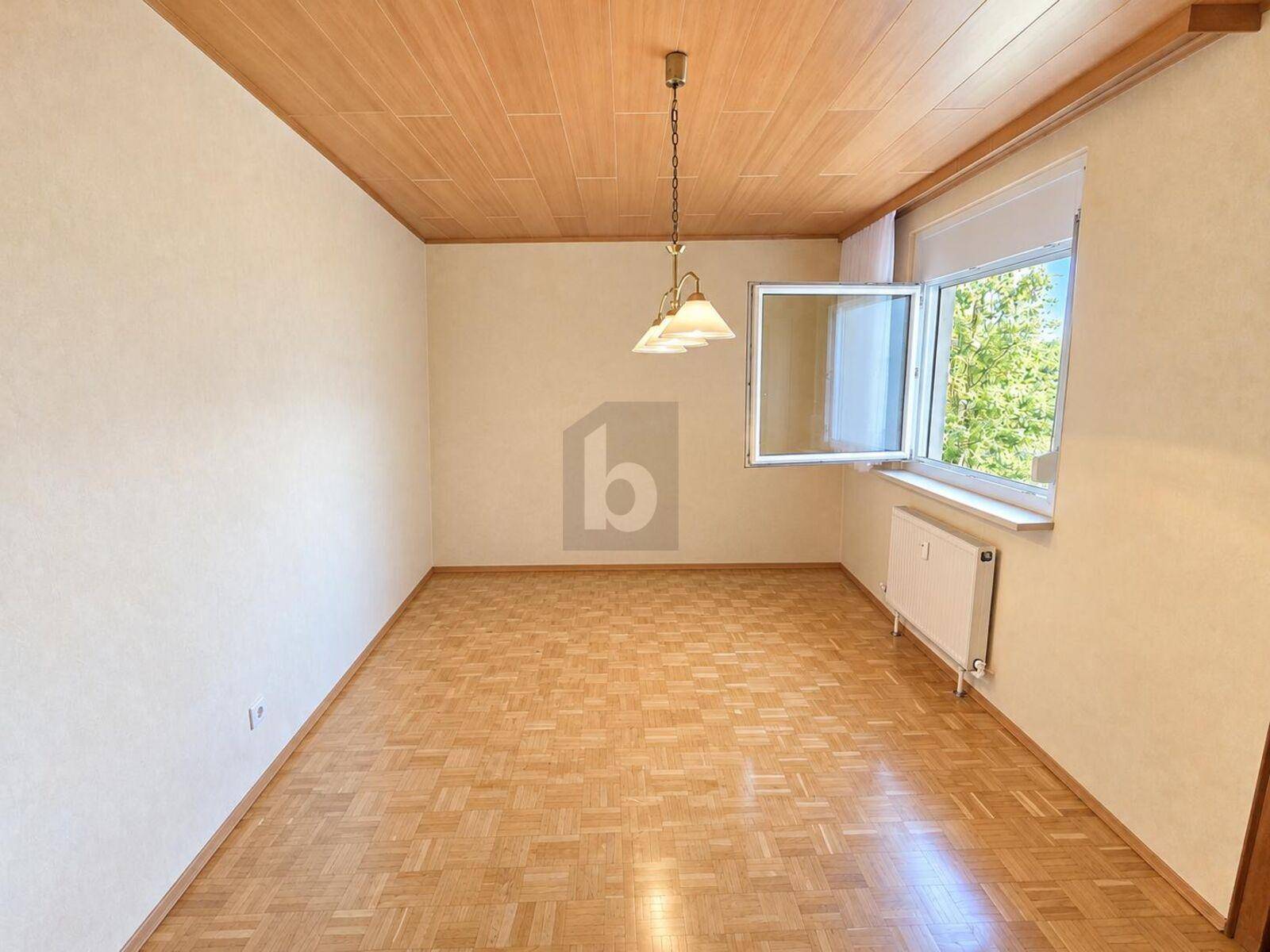 Geräumiger Raum mit Parkettboden, Holzdecke und Fenster mit Blick ins Grüne.