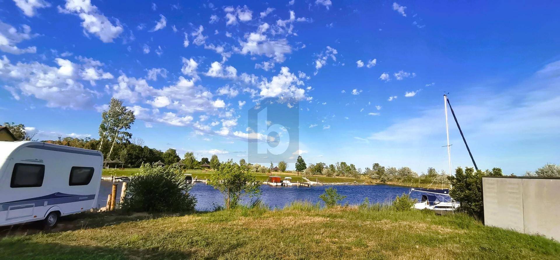 Seeufer mit Segelbooten und einem Wohnwagen, umgeben von grüner Natur und blauem Himmel.