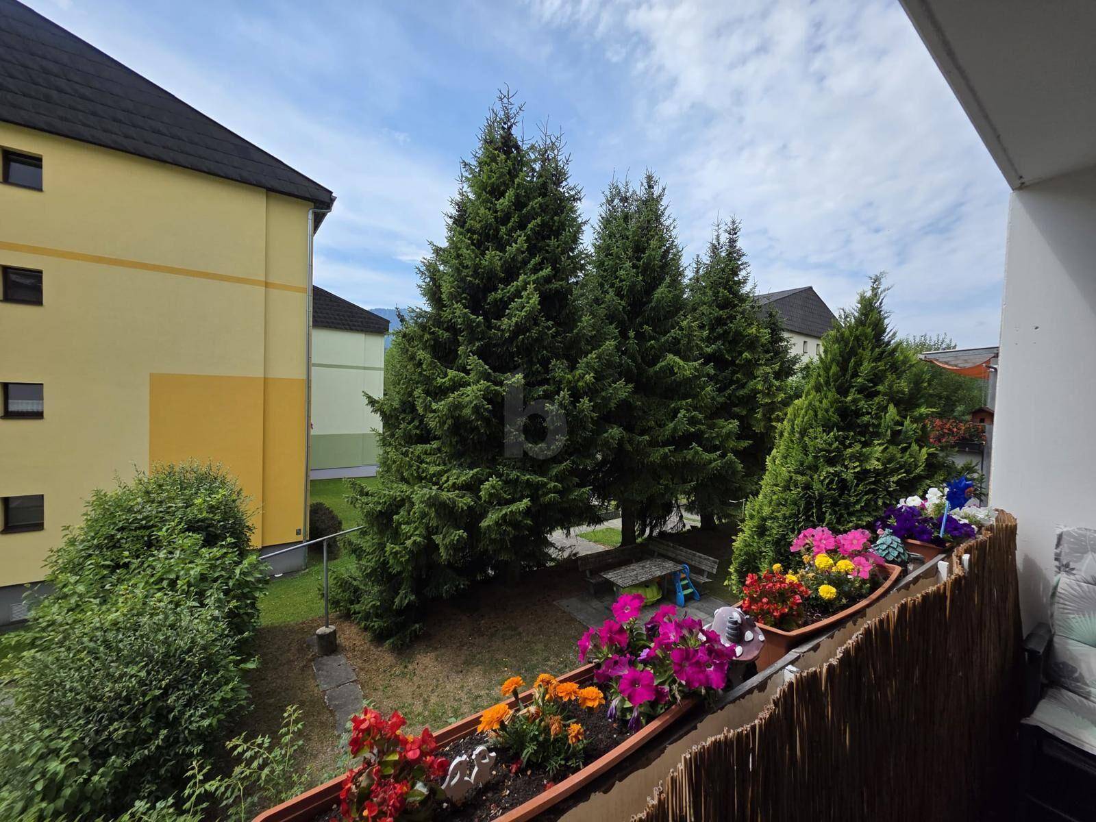 Balkon mit bunten Blumenkästen und Blick auf eine grüne Wohnanlage mit hohen Bäumen.