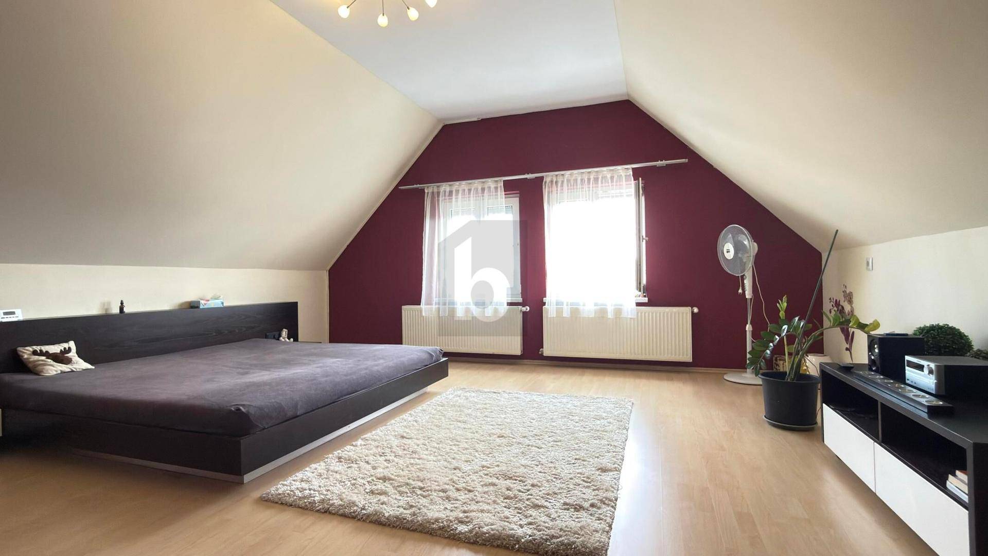 Helles Schlafzimmer im Dachgeschoss mit schrägen Wänden, Laminatboden und einer roten Akzentwand.