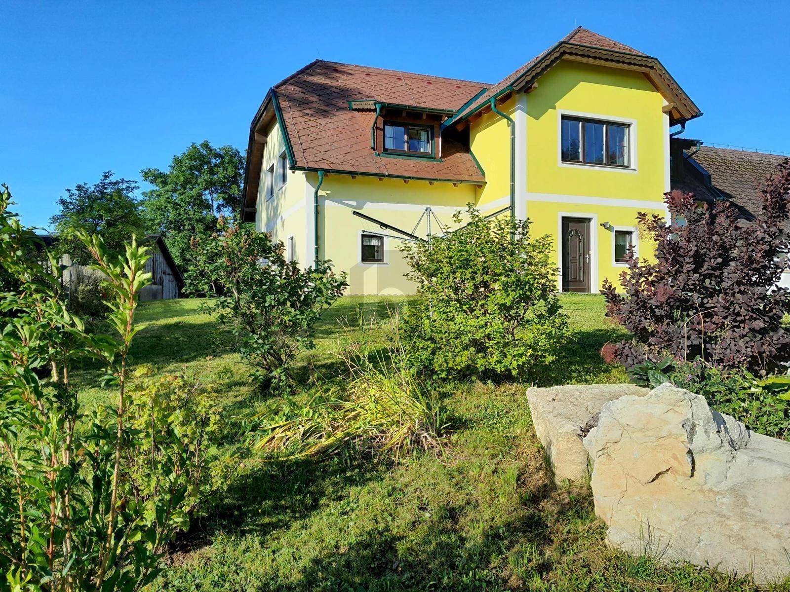 Charmantes gelbes Haus mit braunem Ziegeldach und gepflegtem Gartenbereich an einem sonnigen Tag.