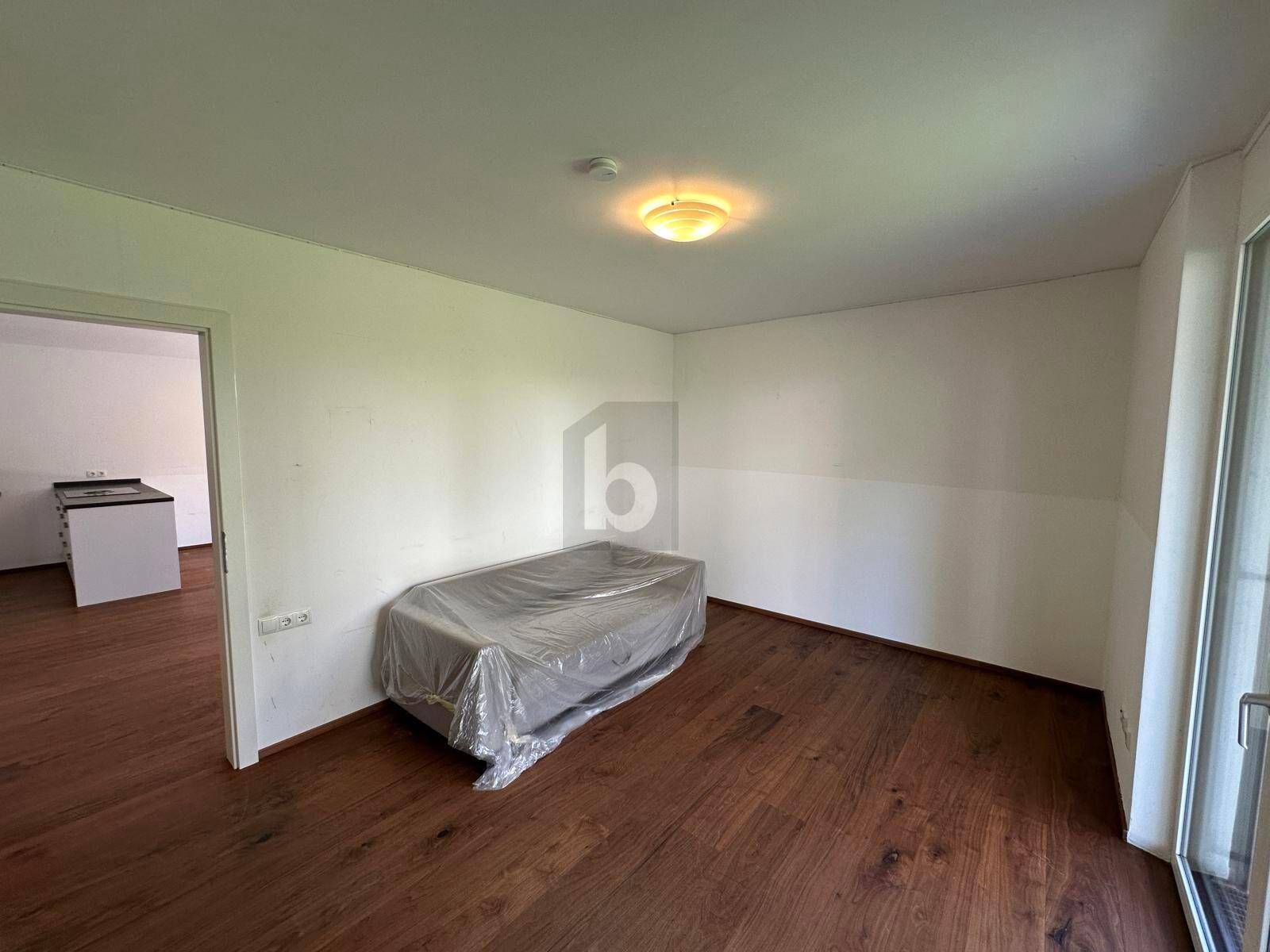 Heller Wohnraum mit Holzboden und Zugang zu einem weiteren Zimmer mit Küchenbereich.