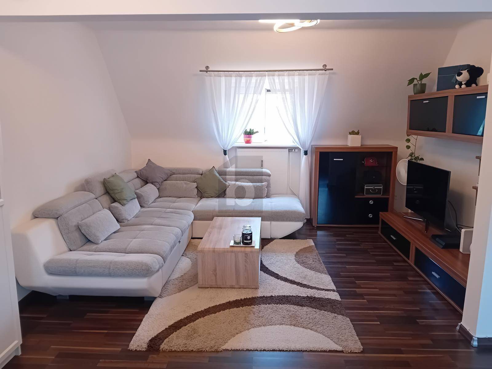Gemütliches Wohnzimmer mit großem Ecksofa, Holzboden und hellem Fenster mit Vorhängen.