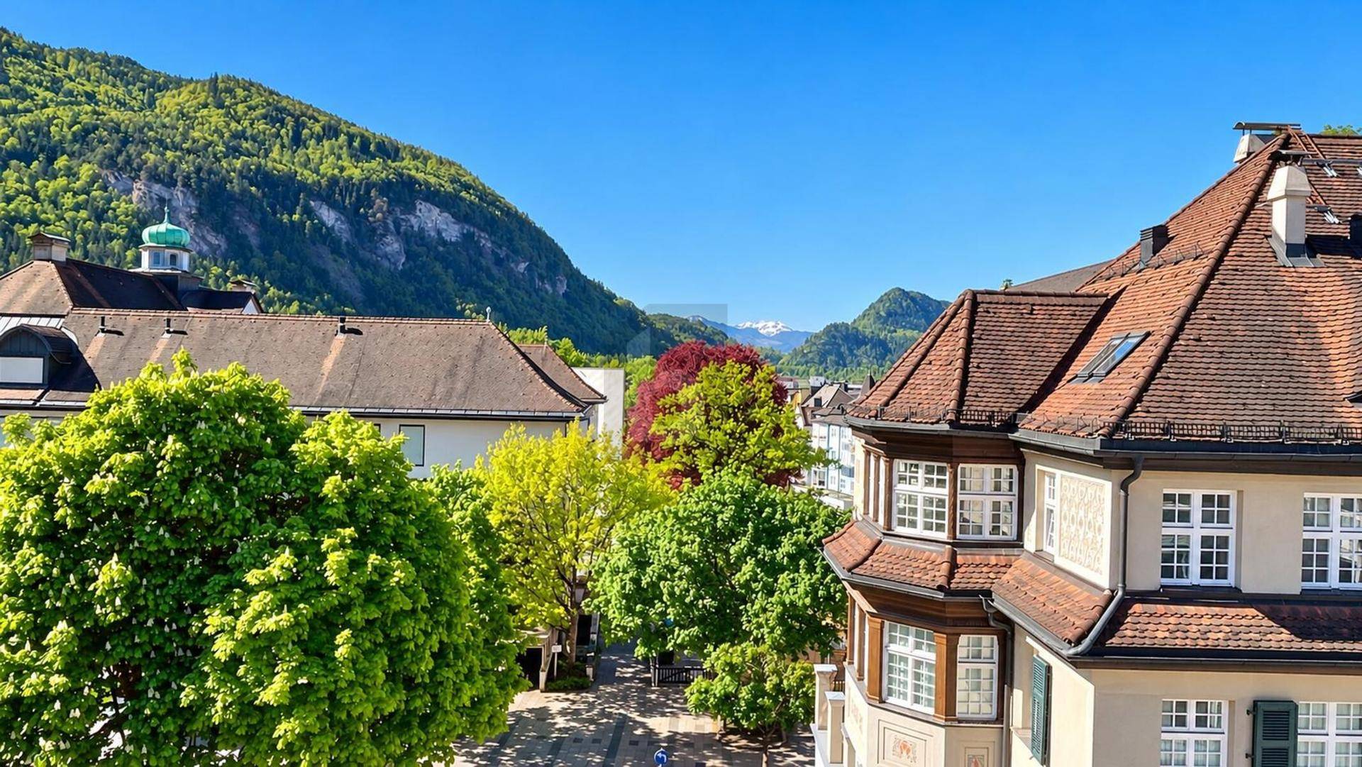 Malerischer Blick über die Dächer der Stadt mit grünen Bergen im Hintergrund unter einem strahlend blauen Himmel.