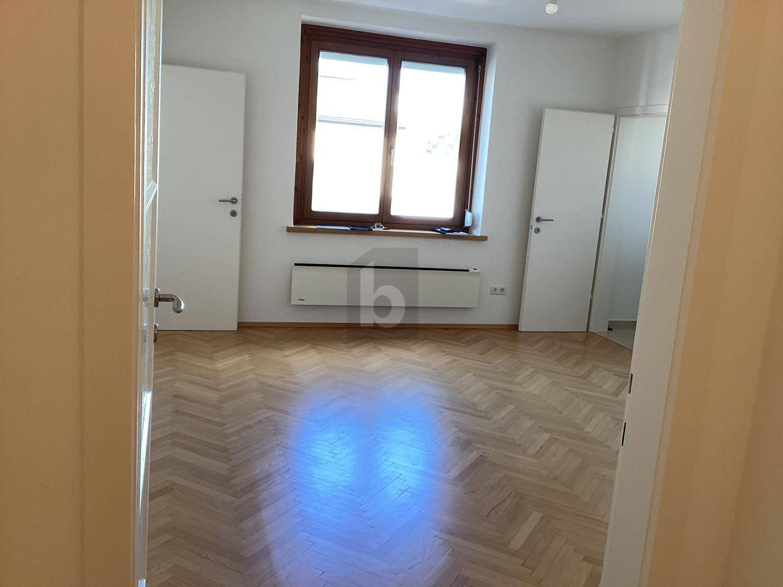 Heller, leerer Raum mit Fischgrätparkettboden, großem Fenster und weißen Türen.