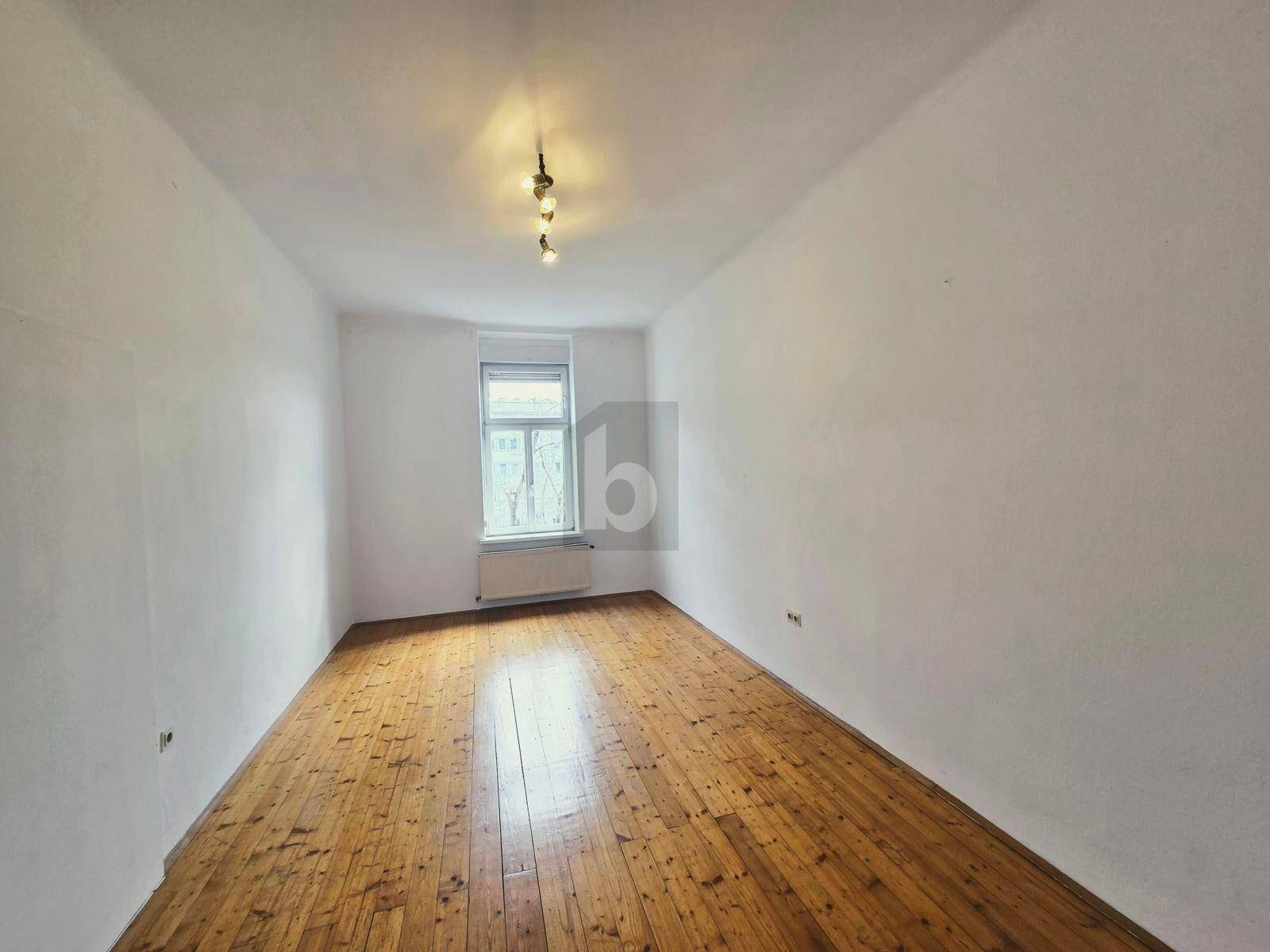 Geräumiges Zimmer mit hellem Holzboden und einem Fenster, das viel Tageslicht hereinlässt.