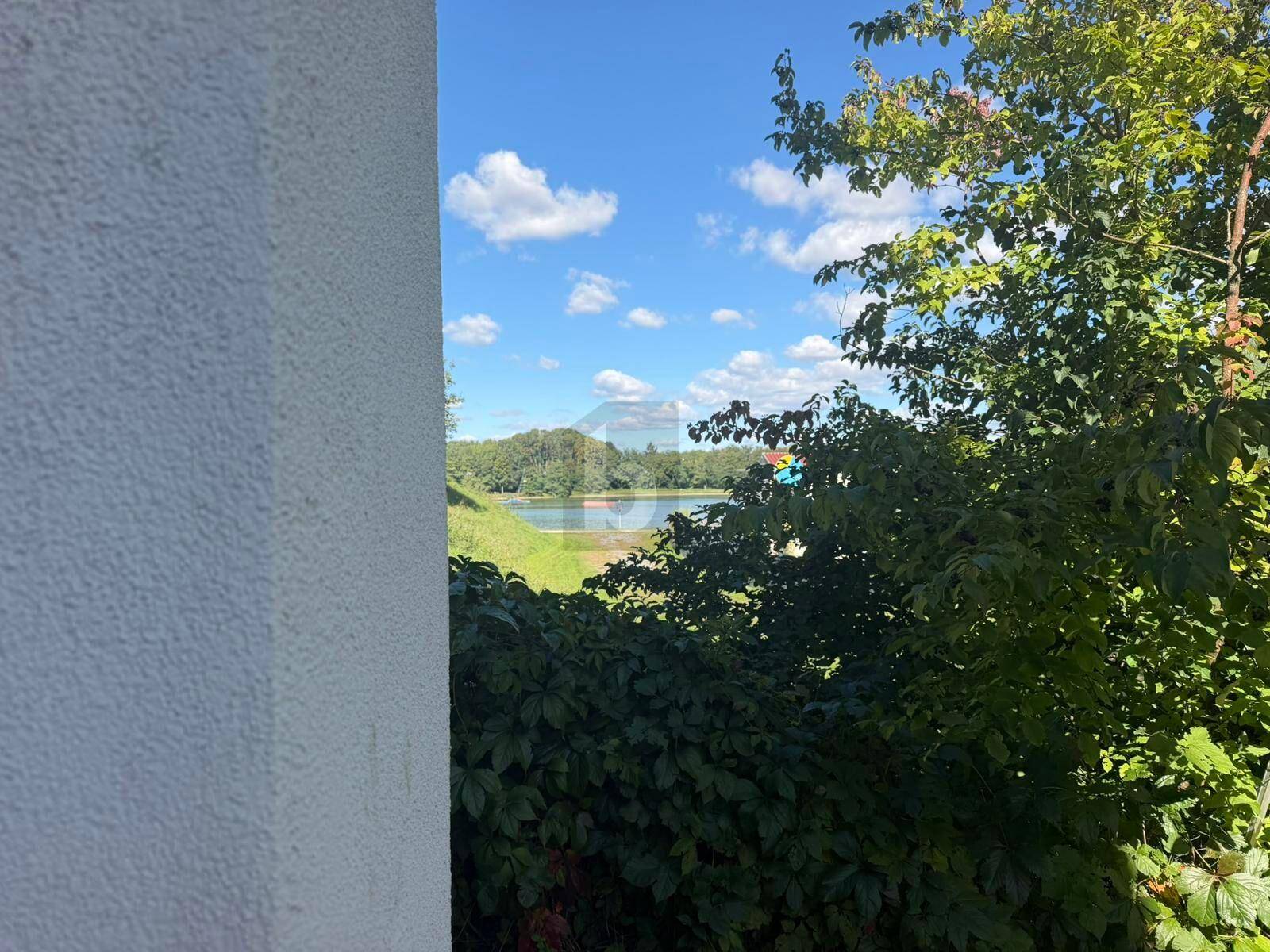 Blick auf einen ruhigen See, umgeben von üppigem Grün und Bäumen unter blauem Himmel.