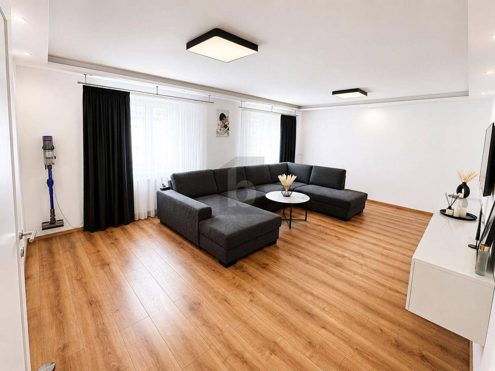 Geräumiges Wohnzimmer mit dunklem Ecksofa, Holzboden und modernen Deckenleuchten.