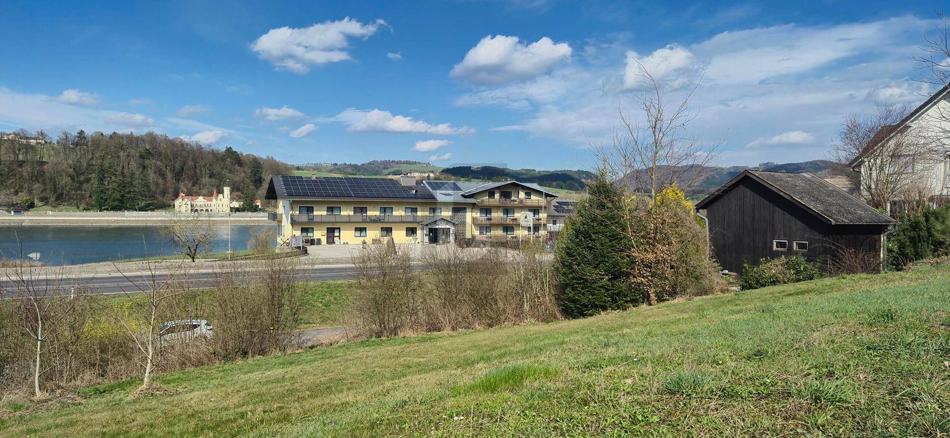 Blick auf ein gelbes Gebäude mit Solaranlage und einen See, umgeben von grüner Landschaft.