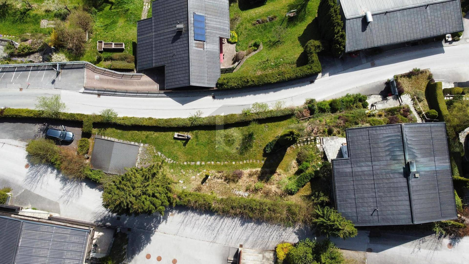 Luftaufnahme einer Wohnsiedlung mit mehreren Häusern, Gärten und Straßen in bergiger Umgebung.