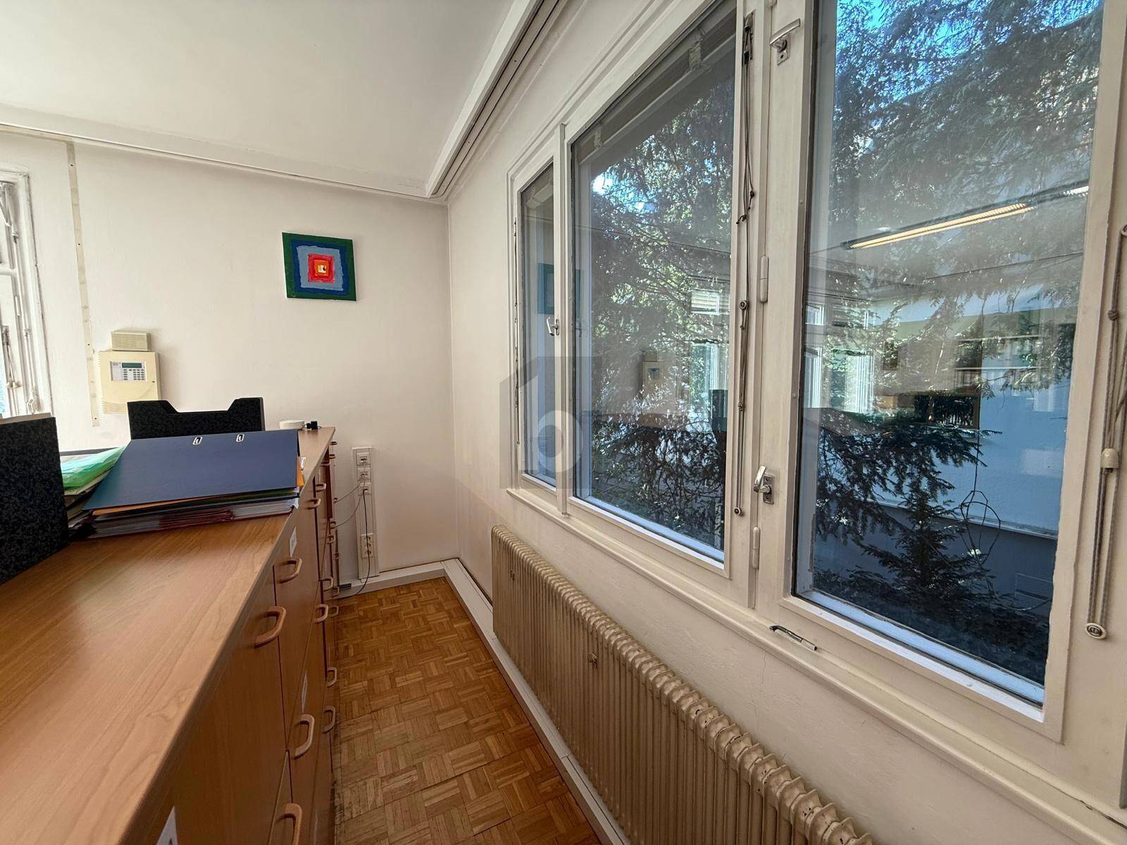 Arbeitsbereich in einem Büro mit Holzparkettboden und großen Fenstern, die einen Blick auf die umliegende Vegetation bieten.