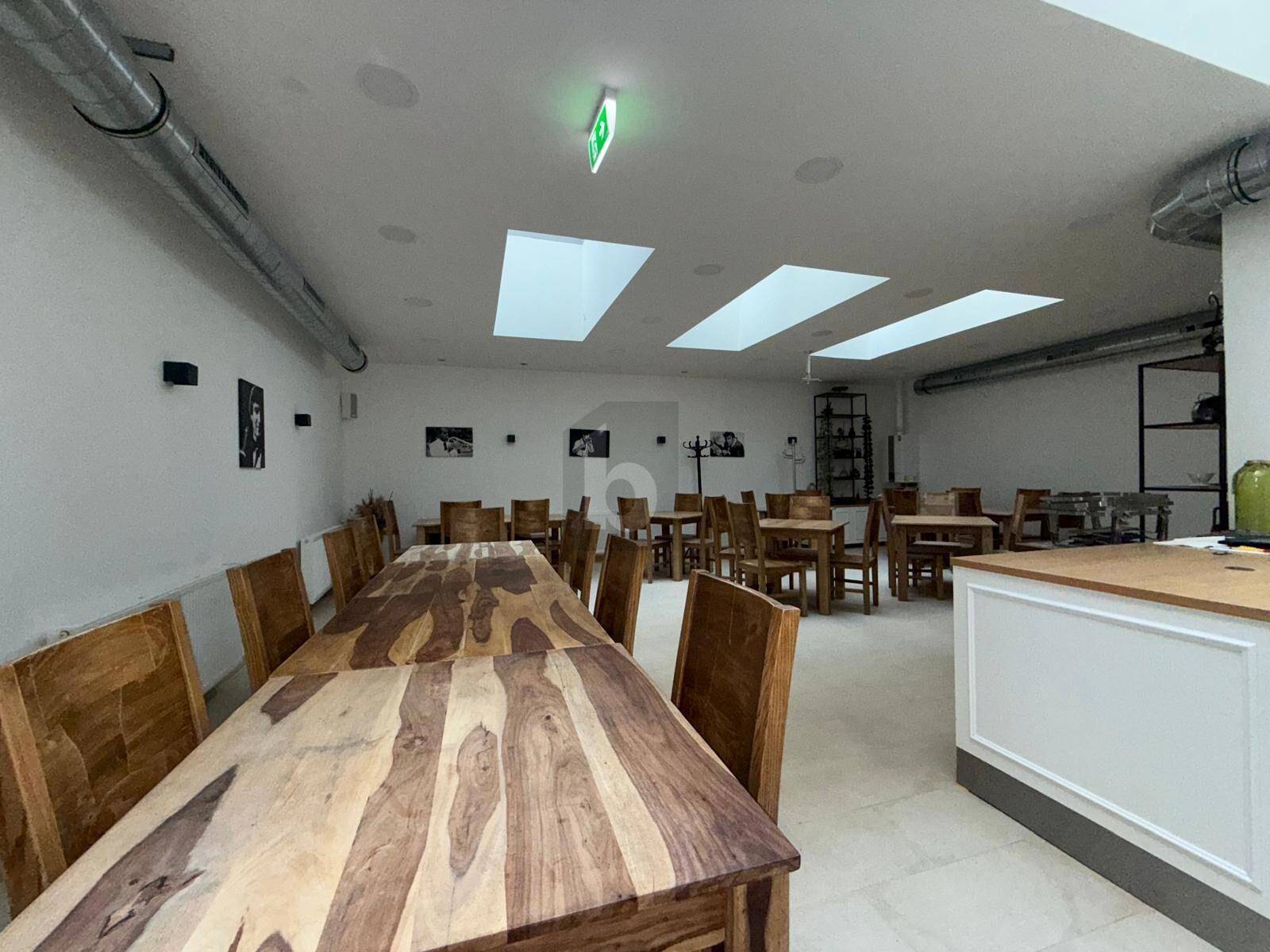 Großer Speisesaal mit langen Holztischen und passenden Stühlen, ergänzt durch Oberlichter und weiße Wände.