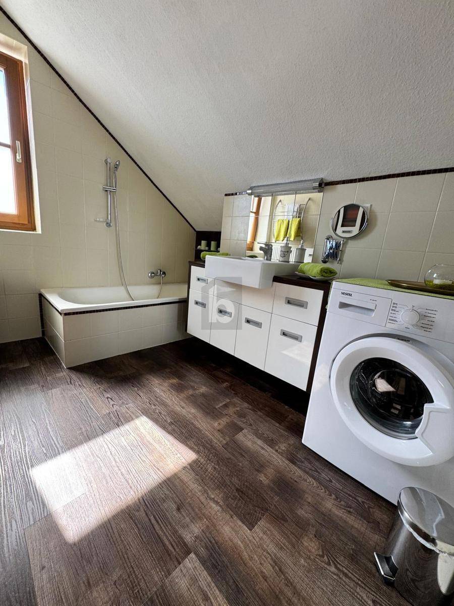 Helles Badezimmer mit Badewanne, Waschbecken und Waschmaschine unter einer Dachschräge.