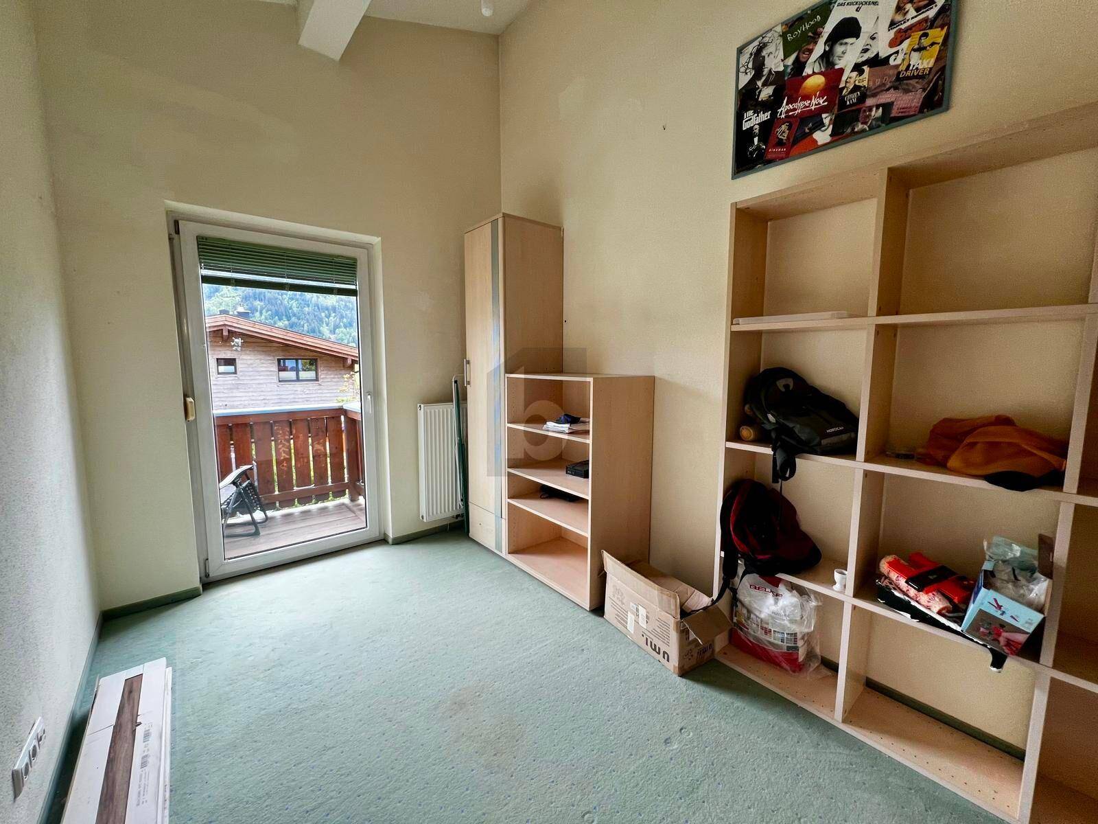 Helles Schlafzimmer mit grünem Teppichboden, Regalen und Zugang zu einem kleinen Balkon.