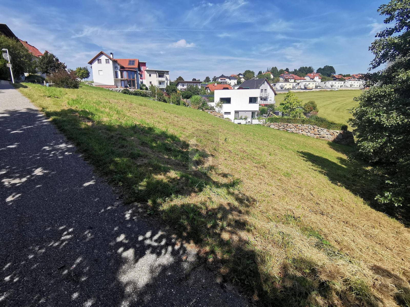 Hanglage mit frisch gemähter Wiese und Blick auf ein modernes Haus sowie weitere Gebäude in der Ferne.