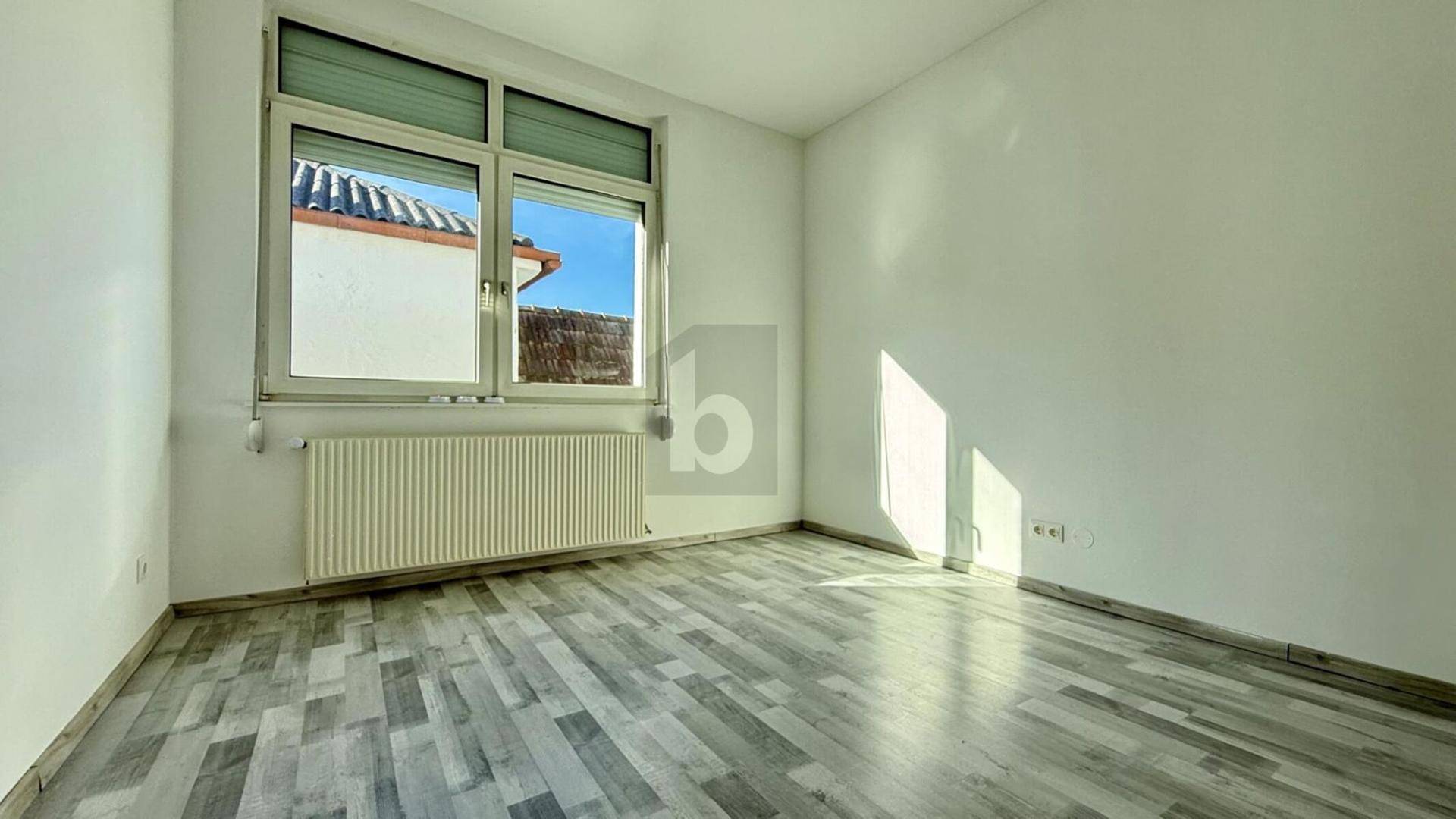 Heller, leerer Raum mit Laminatboden, großem Fenster und Heizkörper.