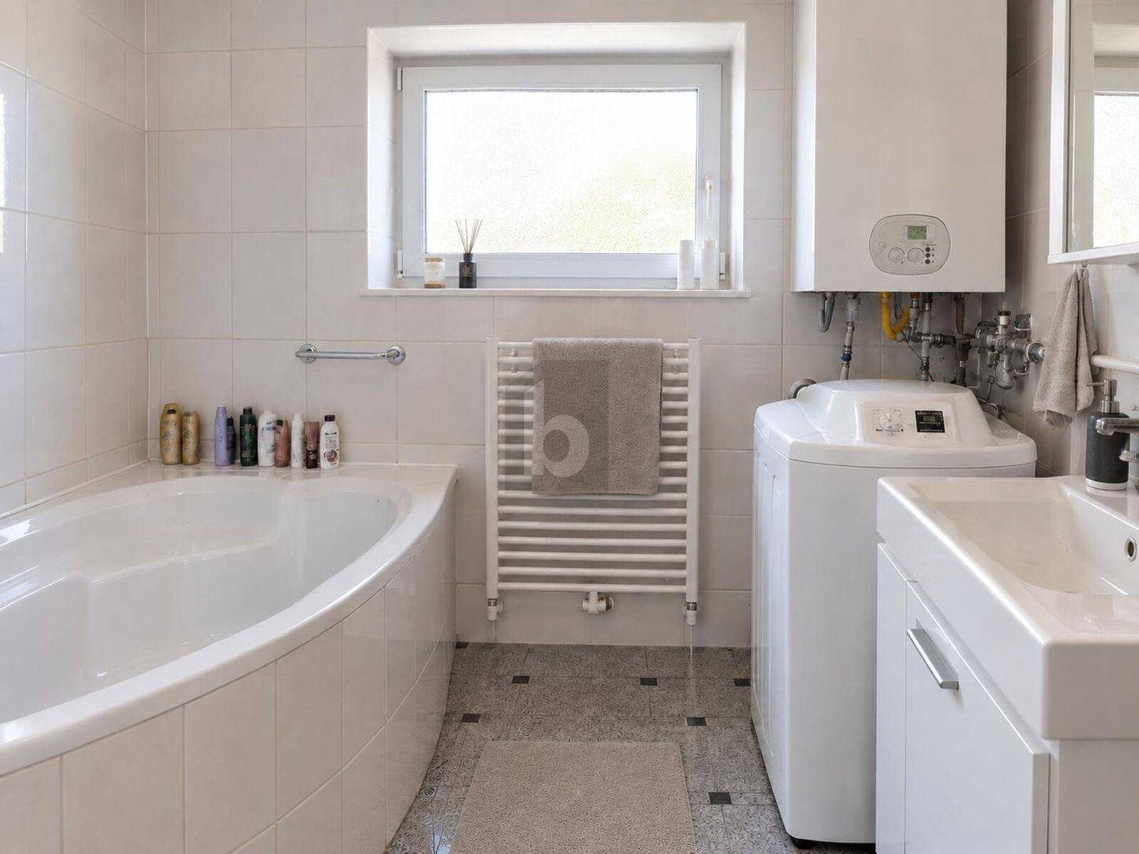 Helles Badezimmer mit Badewanne, Fenster, Handtuchheizkörper und integrierter Heizungsanlage.