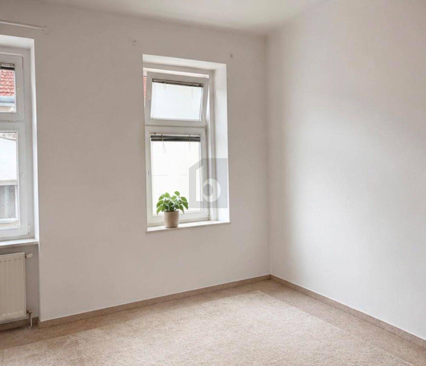 Ein heller, leerer Raum mit weißen Wänden, Fliesenboden und einem Fenster, das viel Tageslicht hereinlässt.