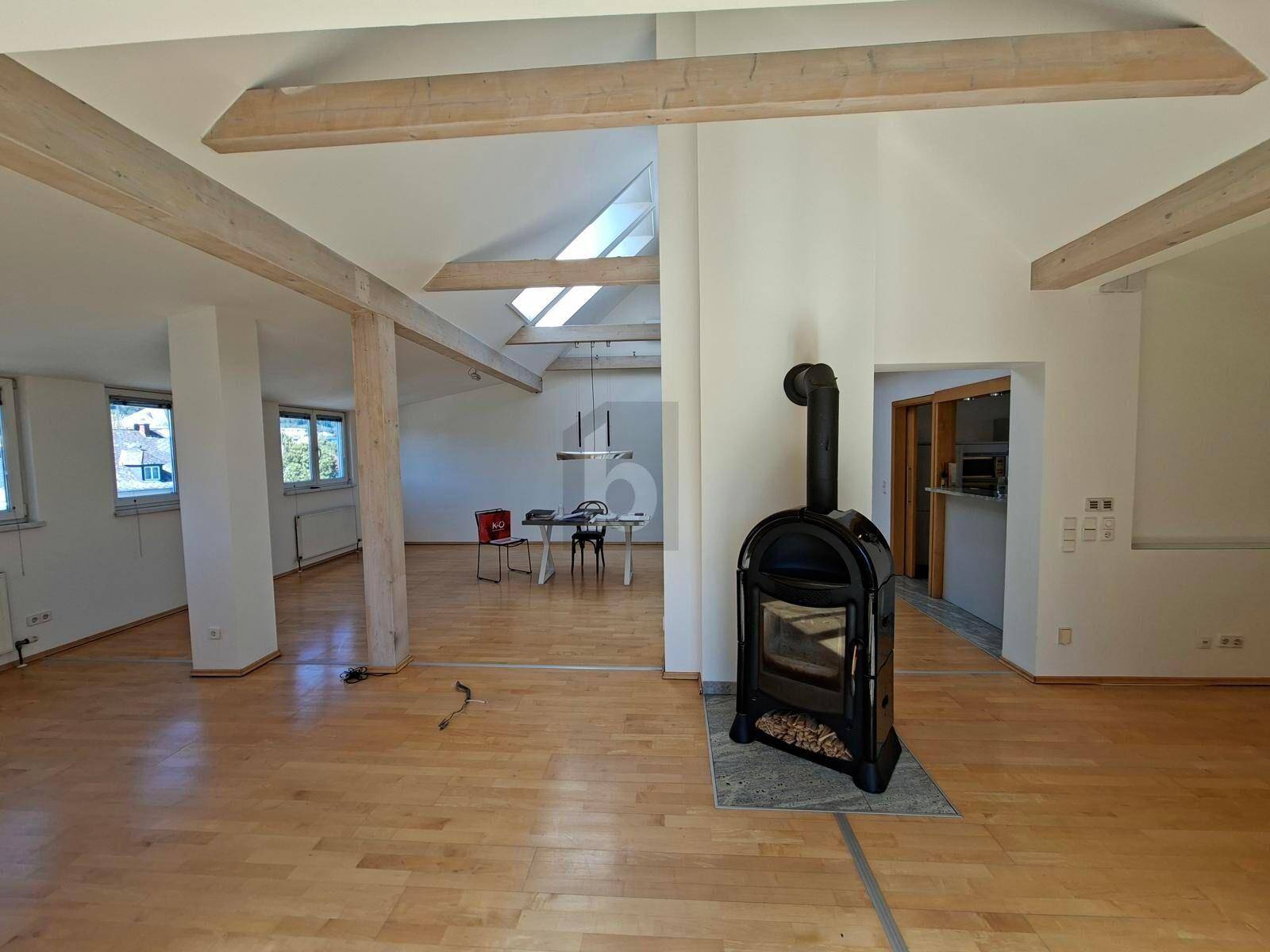 Geräumiger offener Wohnbereich mit Holzboden, sichtbaren Holzbalken und einem modernen Kaminofen.