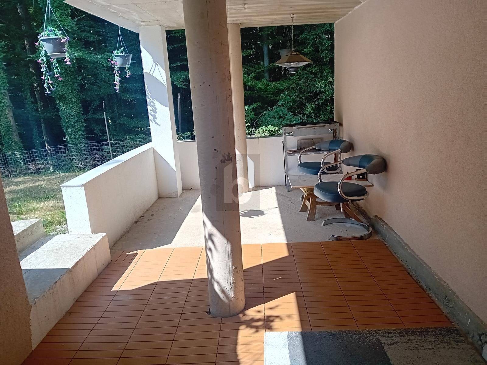Überdachte Terrasse mit braunen Fliesen und Sitzgelegenheiten, umgeben von üppigem Grün.
