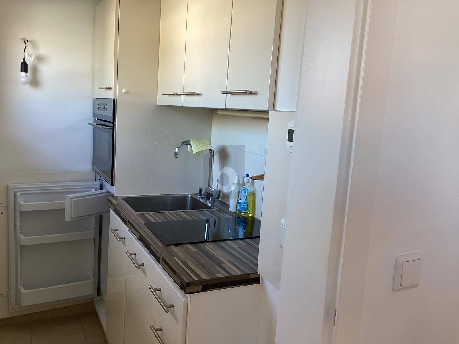 Kompakte Einbauküche mit weißen Schränken, Spüle, Kochfeld, Backofen und kleinem Kühlschrank.