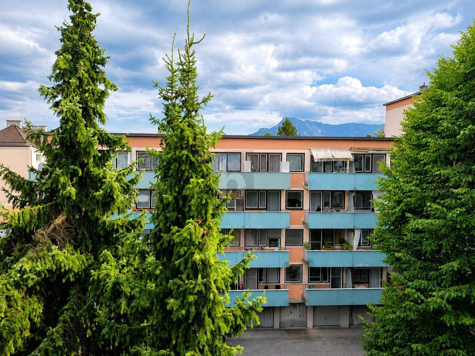 Mehrfamilienhaus mit Balkonen, umgeben von Bäumen und Bergen im Hintergrund.