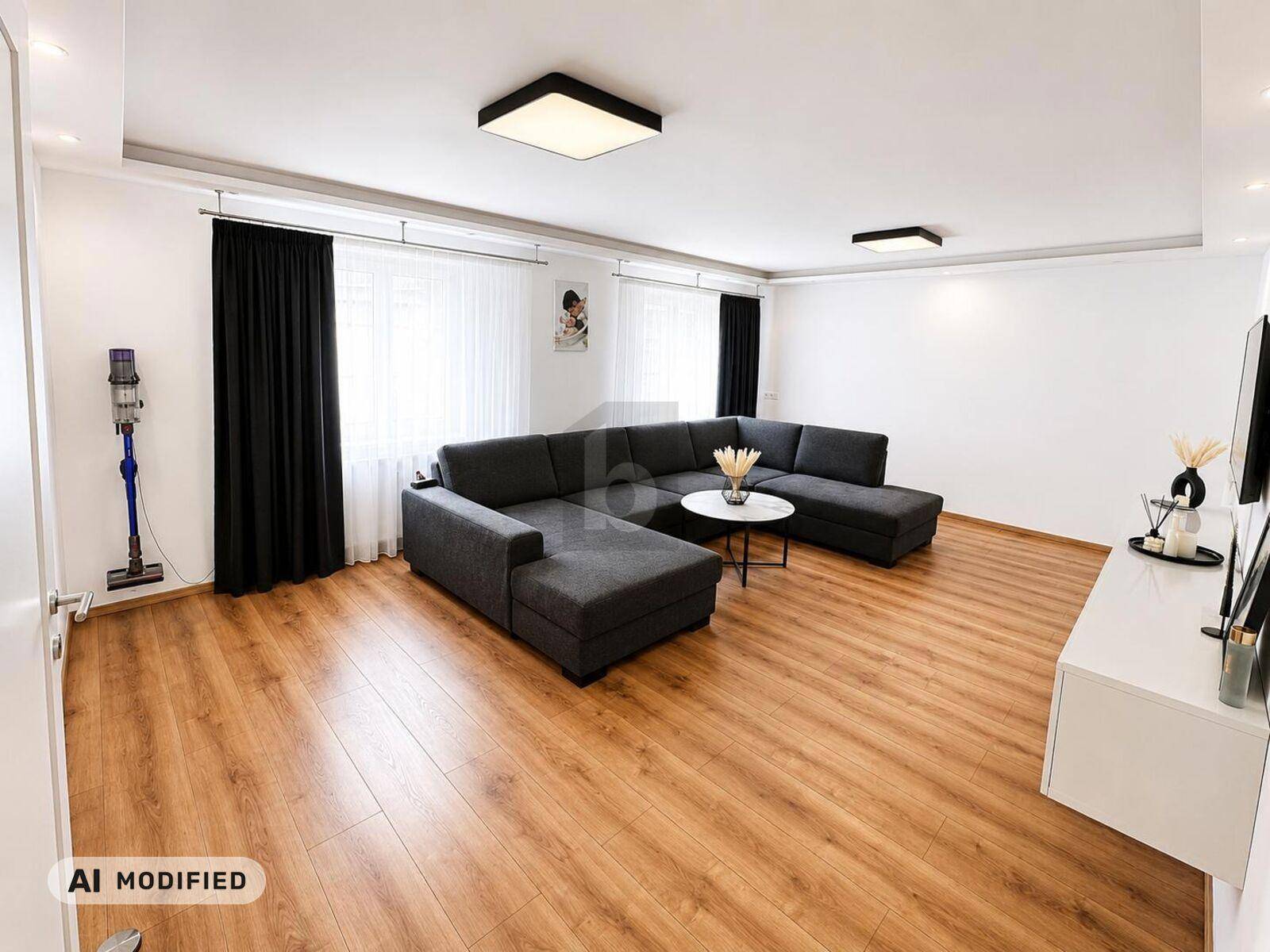 Geräumiges Wohnzimmer mit dunklem Ecksofa, Holzboden und großen Fenstern für viel Tageslicht.