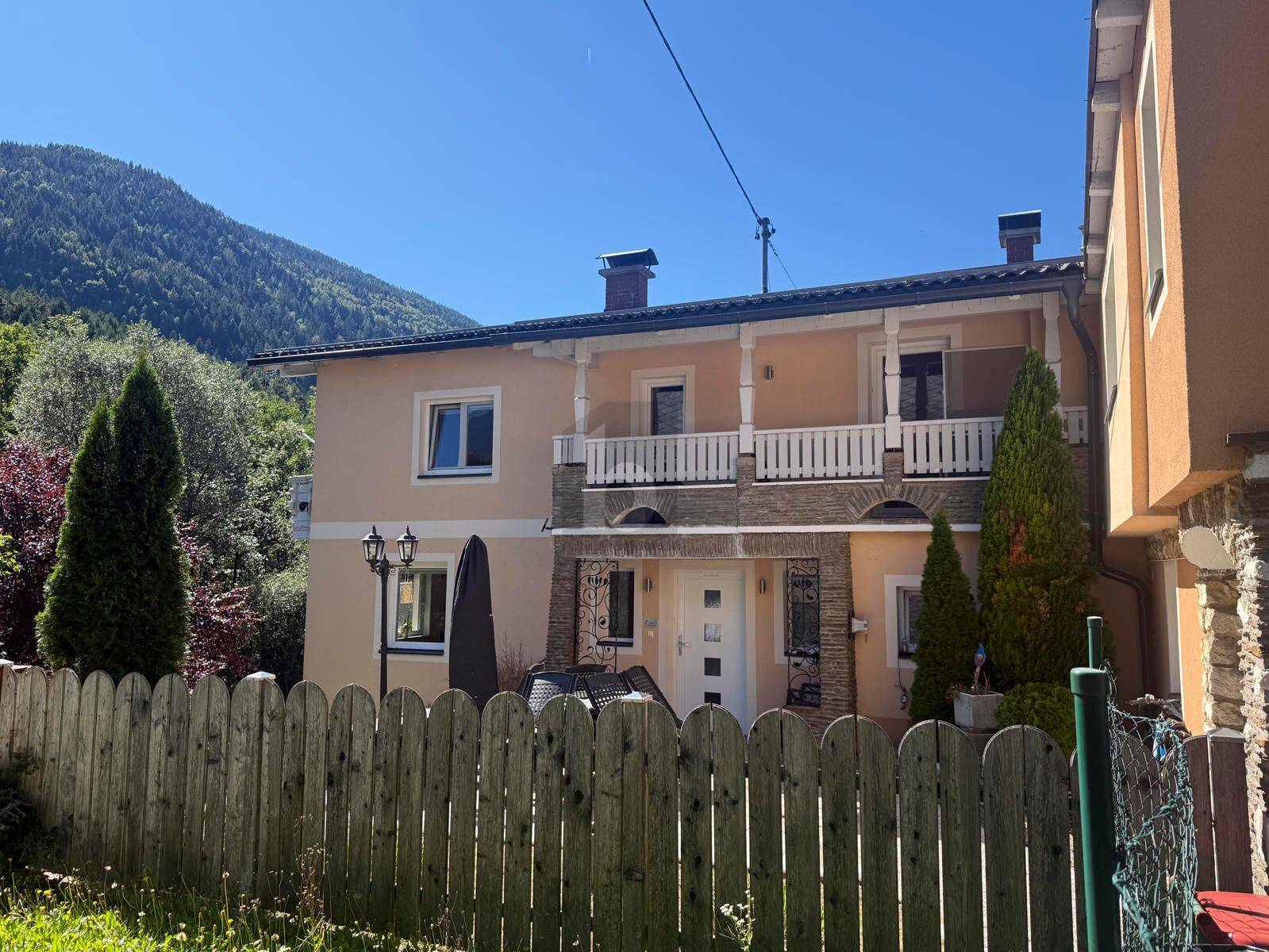 Zweistöckiges Haus mit hellem Anstrich, Balkon und Holzzaun im Vordergrund, umgeben von Bergen.