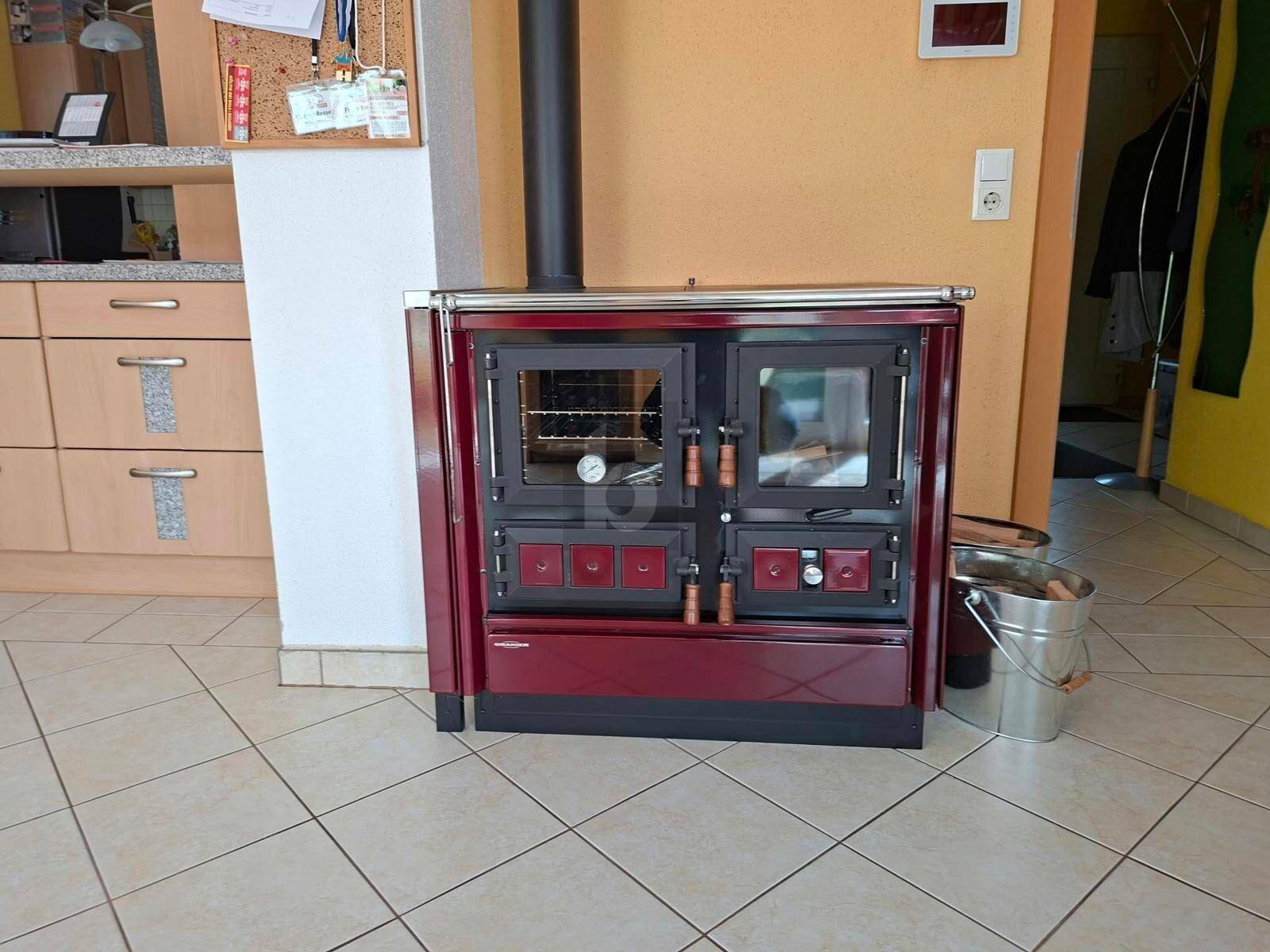 Traditioneller roter Holzofen mit Backfach und Kochplatte auf Fliesenboden.