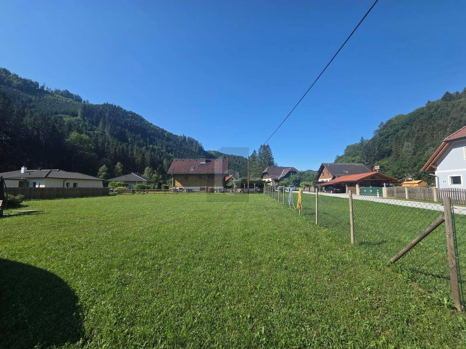 Grüne Wiese mit Zaun im Vordergrund, umgeben von Häusern und bewaldeten Hügeln unter klarem Himmel.